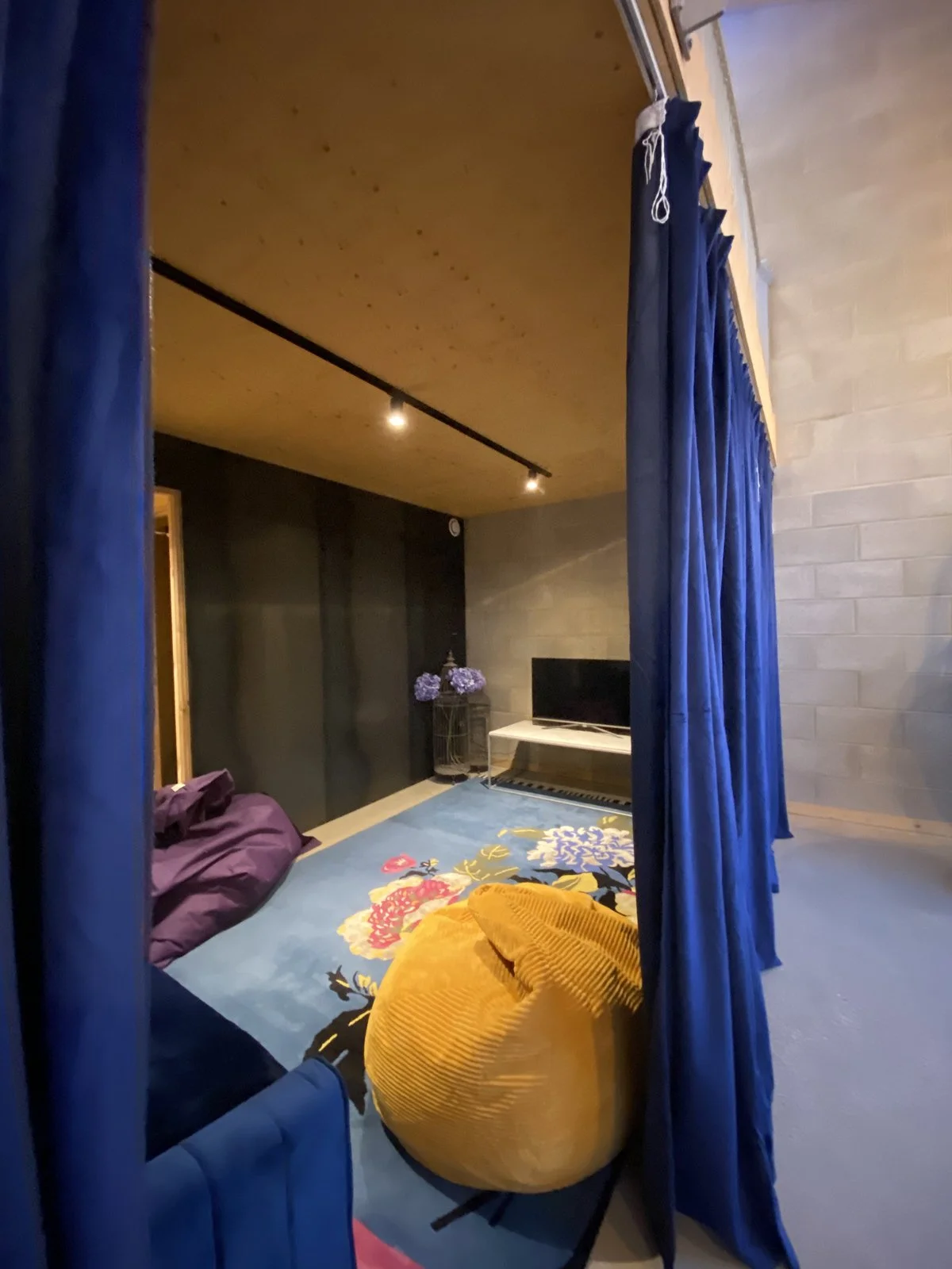Cosy bedroom with blue curtains, purple and yellow pillows, a floral area rug, a small television, and a flower arrangement on a stand.