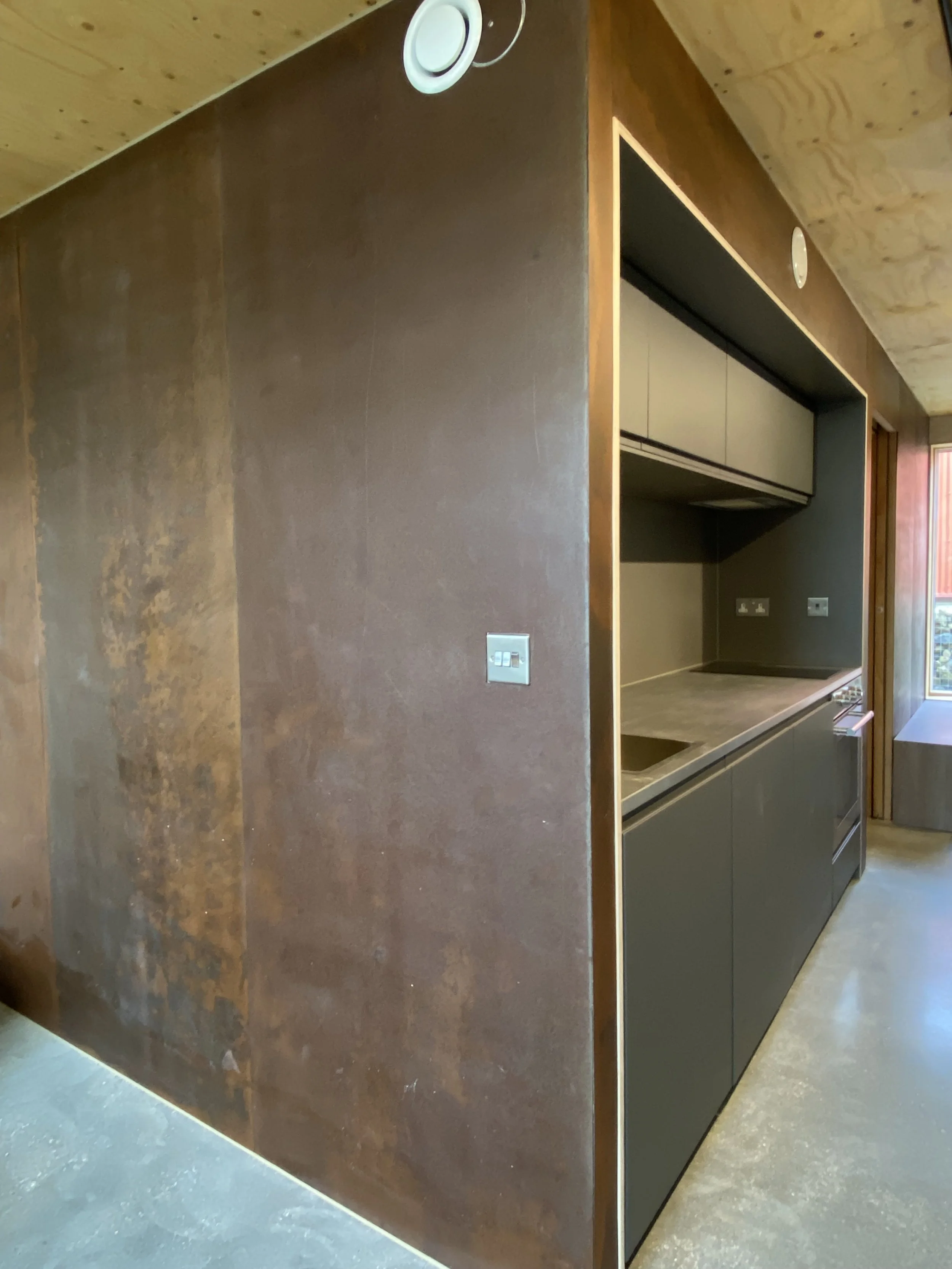A modern kitchen with dark lower cabinets, a built-in countertop, and upper cabinets inside a recessed area. The walls and ceiling are made of wood. Multiple electrical outlets are visible on the wall.