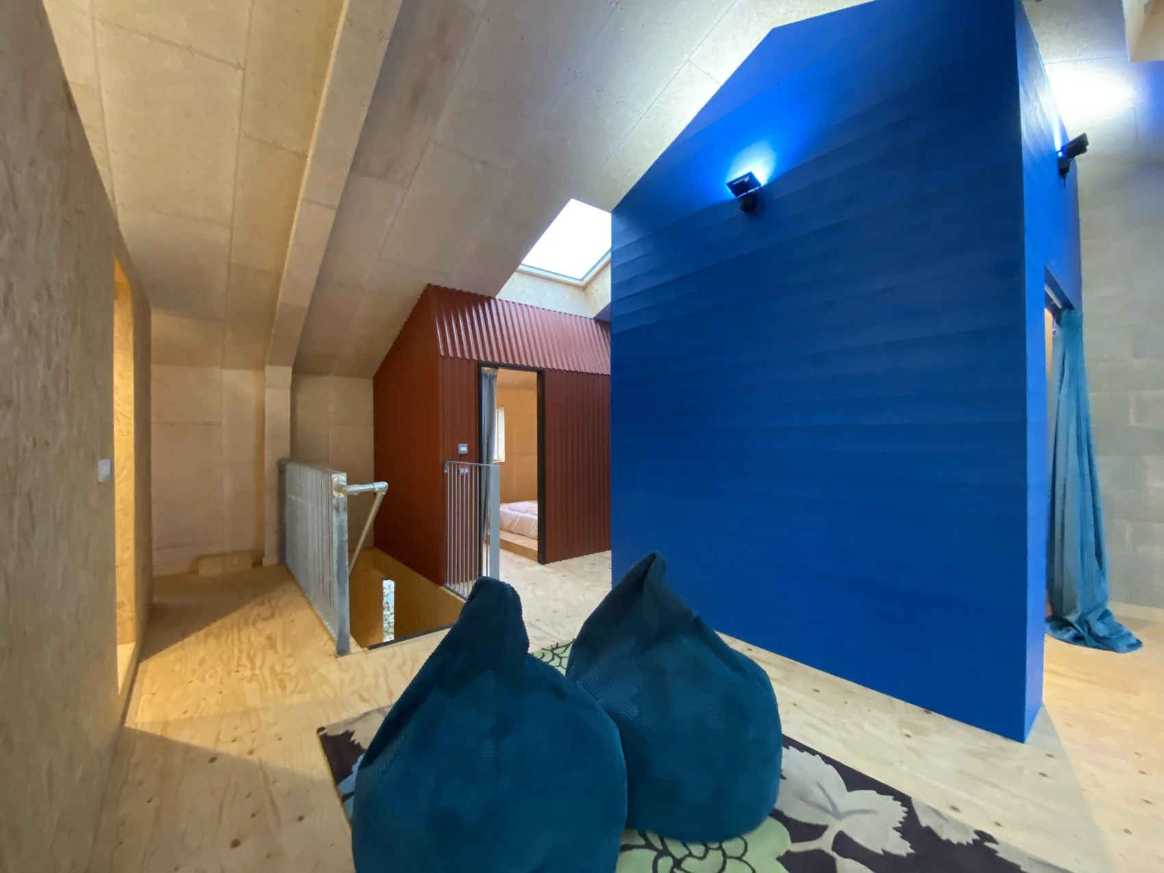 Modern interior space with wooden floors, a bright blue accent wall, and a red partition leading to a bedroom. Two blue bean bag chairs sit on a patterned rug.