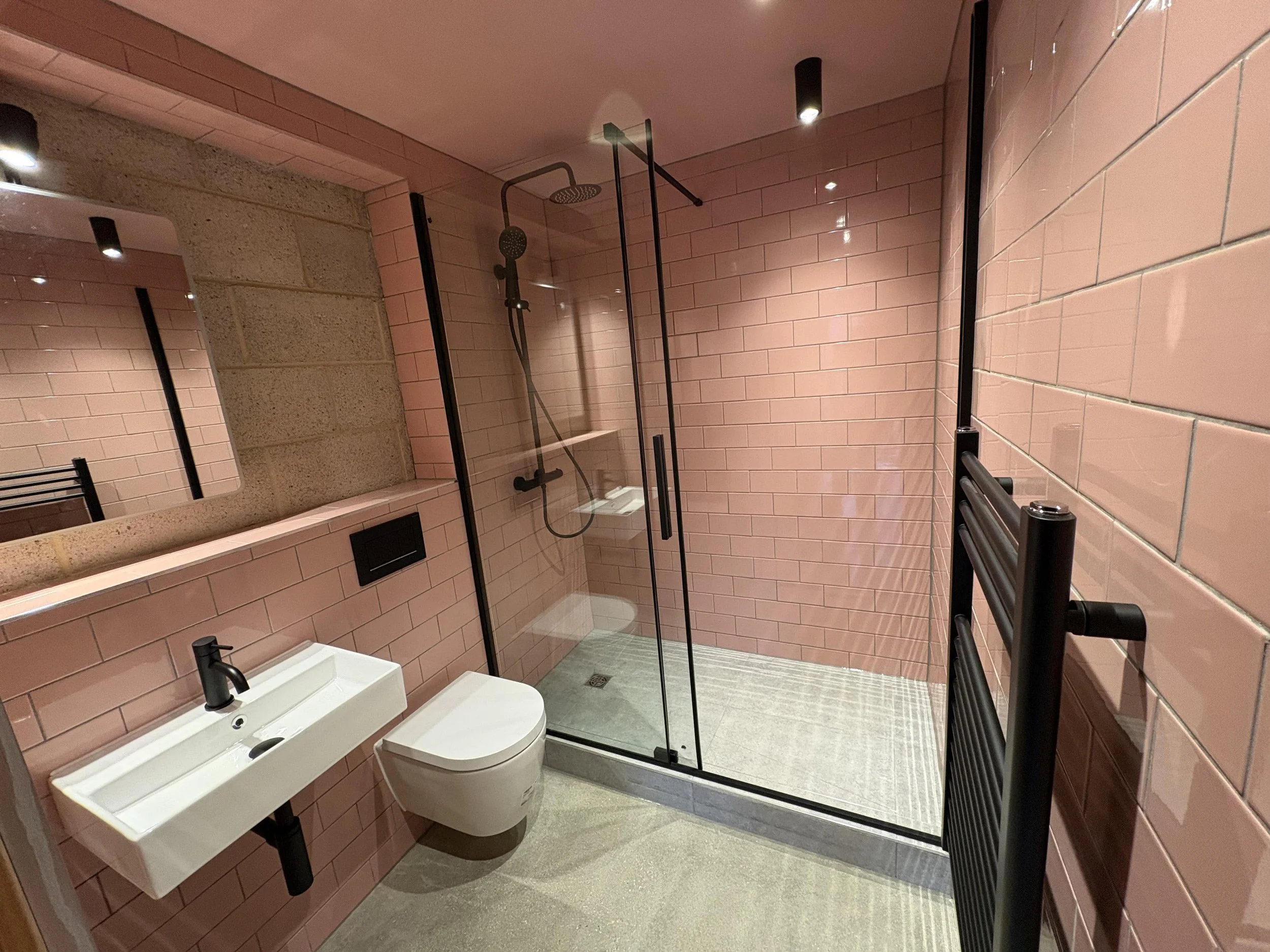 Modern bathroom with pink tiled walls, a glass shower enclosure, a white wall-mounted sink with a black faucet, a wall-mounted toilet, a large rectangular mirror, a black towel rail, and grey flooring.