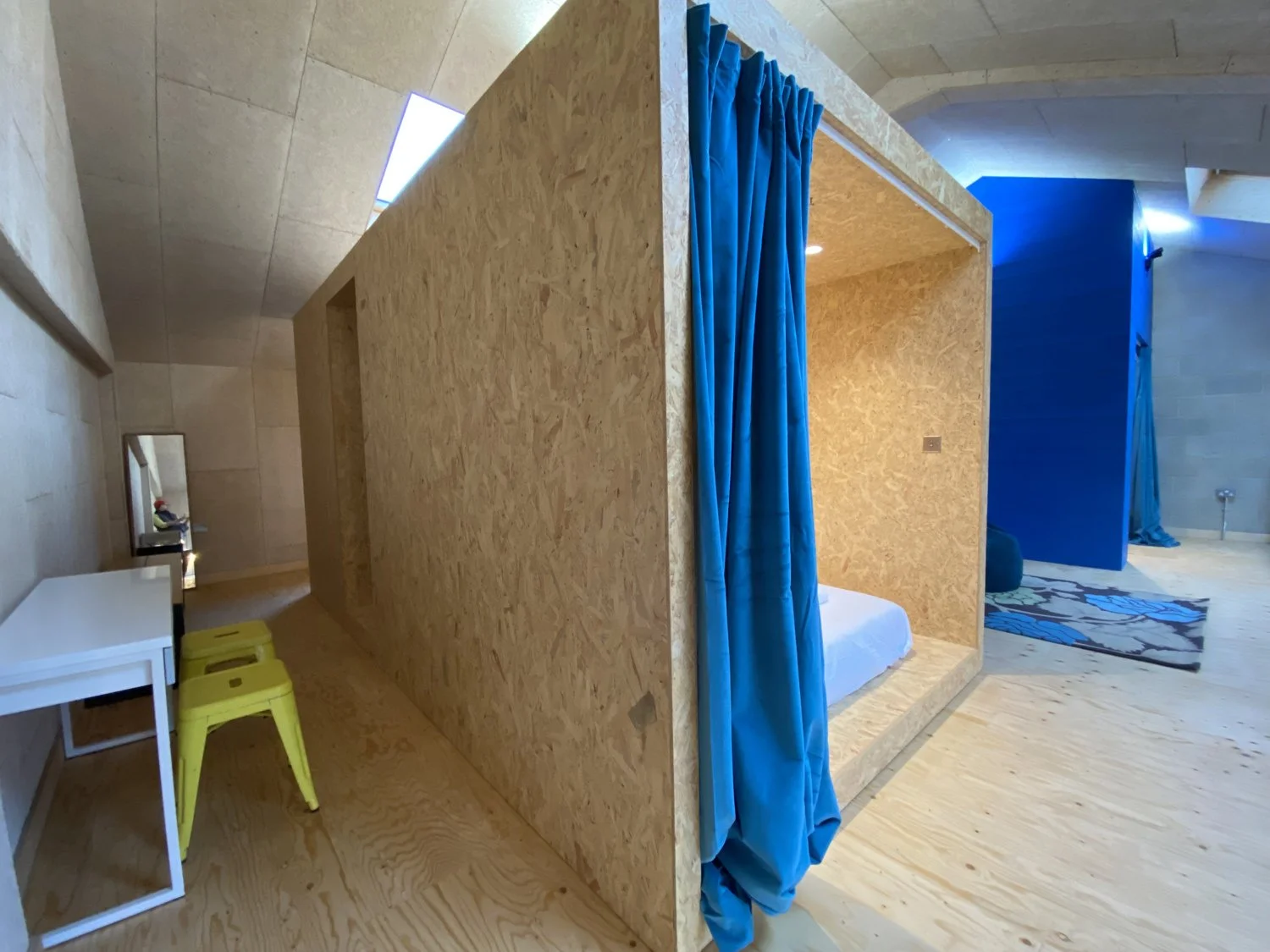 Interior of a modern, minimalistic space with a small wooden house or pod featuring an open front draped with blue curtains, a bed inside, a white desk with yellow stools, and a blue wall with a small rug, wood flooring, and geometric ceiling design.