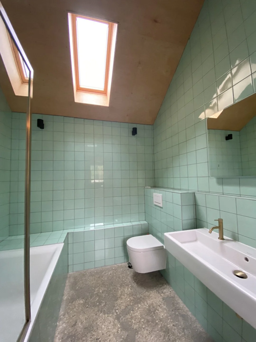 Bathroom with light green tiled walls, skylights in wooden ceiling, a white wall-mounted toilet, a white rectangular sink with a gold faucet, a mirror, and a partial shower area with glass door.
