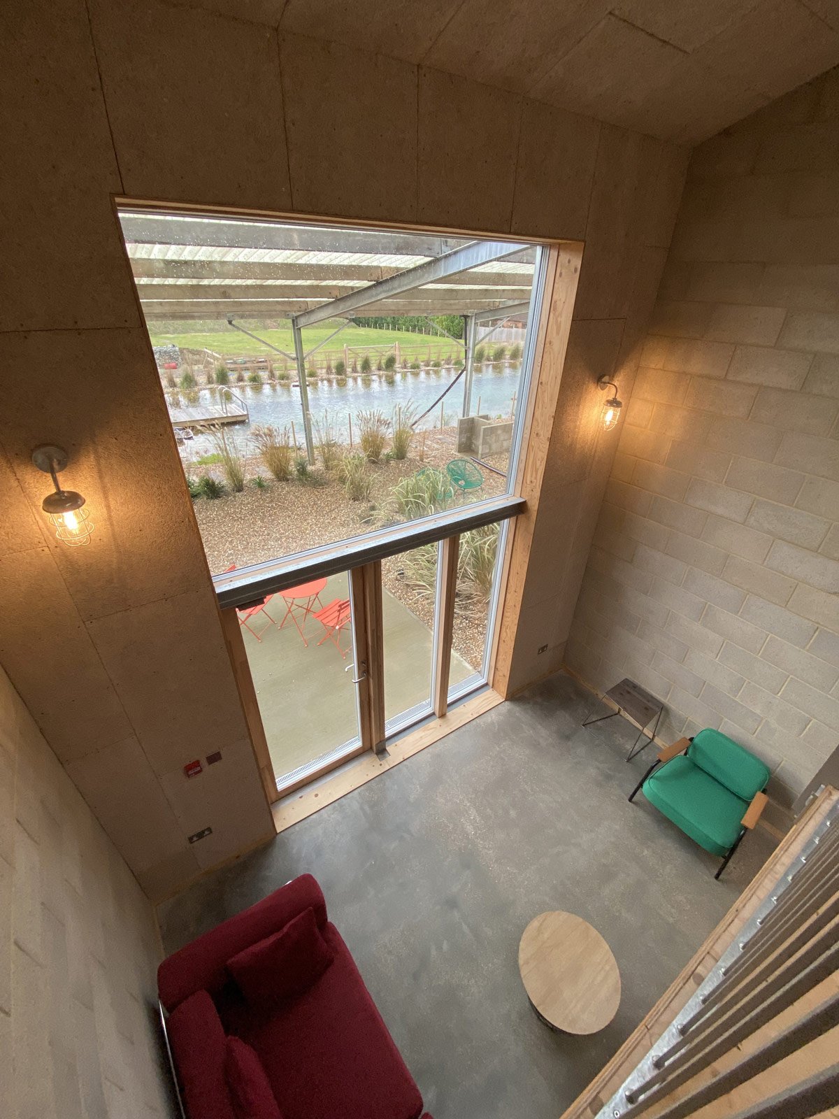 Interior view of a room with a large window showing an outdoor patio and a pond. The room has a red sofa, a green armchair, a small side table, and a round wooden coffee table. The walls are made of brick and wood, and there are two wall-mounted ligh