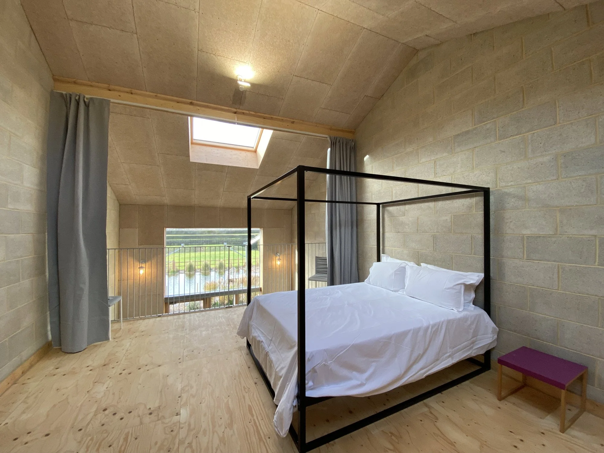 Minimalist bedroom with a black bed frame, white bedding, and natural wood flooring. Features a skylight and large window overlooking greenery, with gray curtains and a small purple bench.