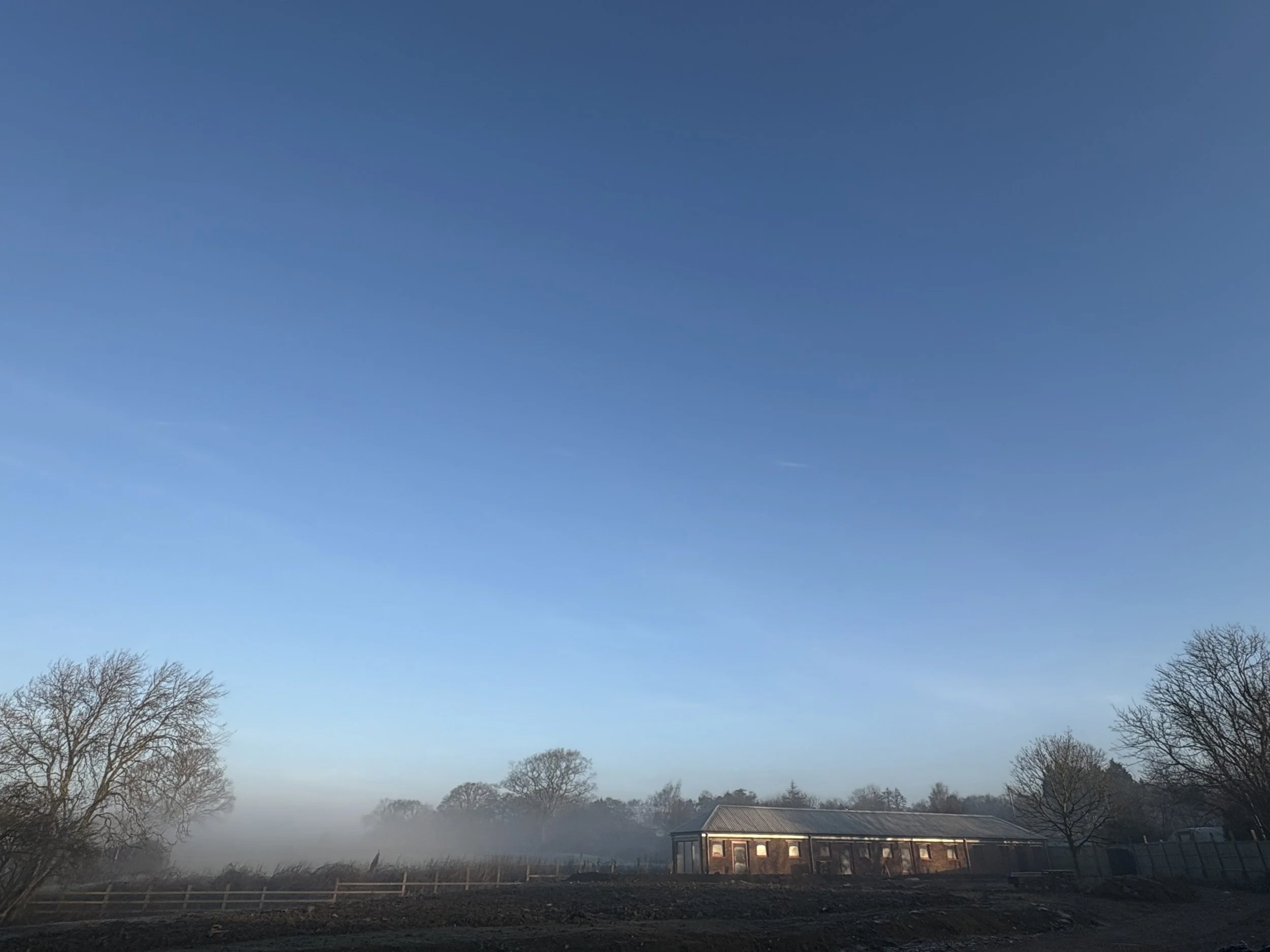 A rural scene with a clear blue sky, leafless trees, a misty horizon, and a long, low building with a metal roof and multiple windows.