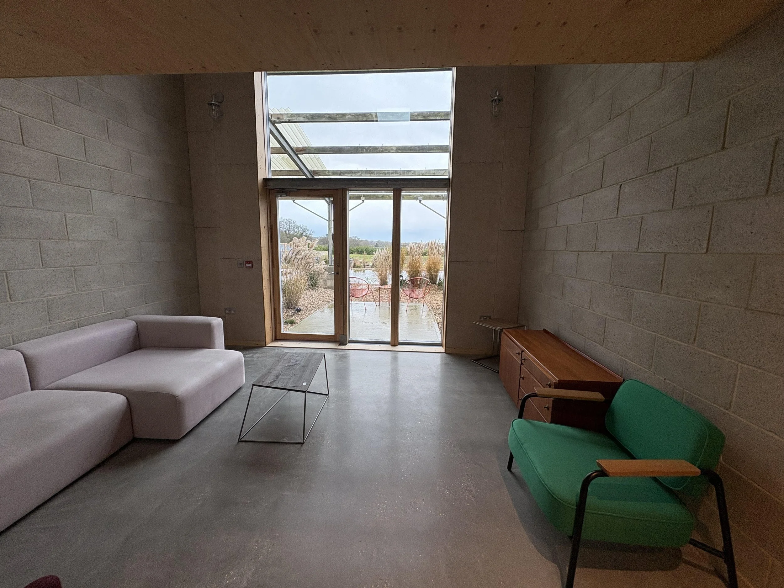 Modern living room with white sofa, green armchair, wooden sideboard, and floor-to-ceiling glass door leading outside to patio with outdoor chairs, overlooking a landscaped yard with ornamental grasses.