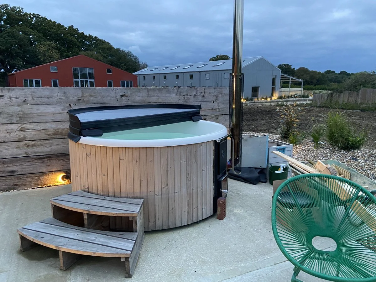 An outdoor hot tub with a wooden exterior, situated on a concrete patio, next to a wooden privacy fence and facing a landscape with a red and a gray building in the background.
