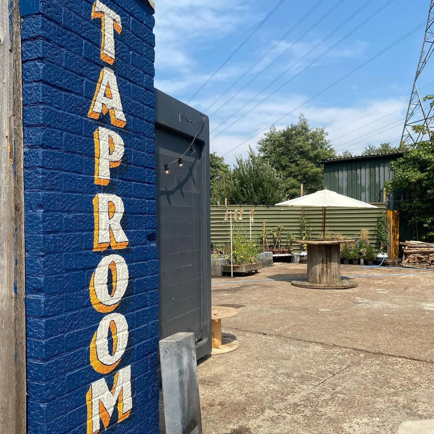 A blue brick wall with the word "TAPROOM" painted in white and yellow letters. Outside, there are wooden tables, an umbrella, plants, and a fence under a partly cloudy sky.