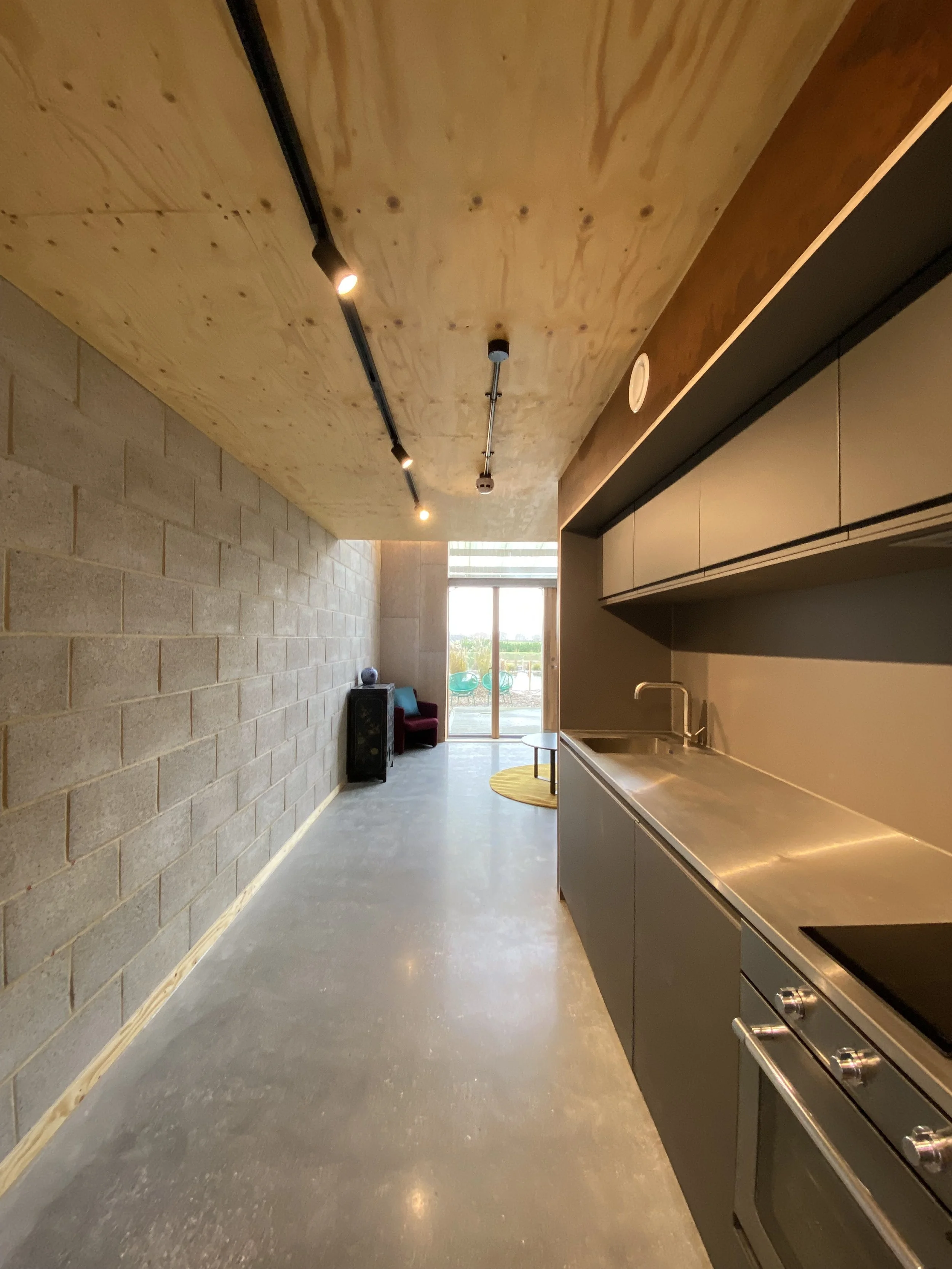Modern minimalist kitchen with stainless steel countertop and cabinets, gray floors, exposed wooden ceiling, brick wall, and a view of a living area with a sliding glass door leading outside.