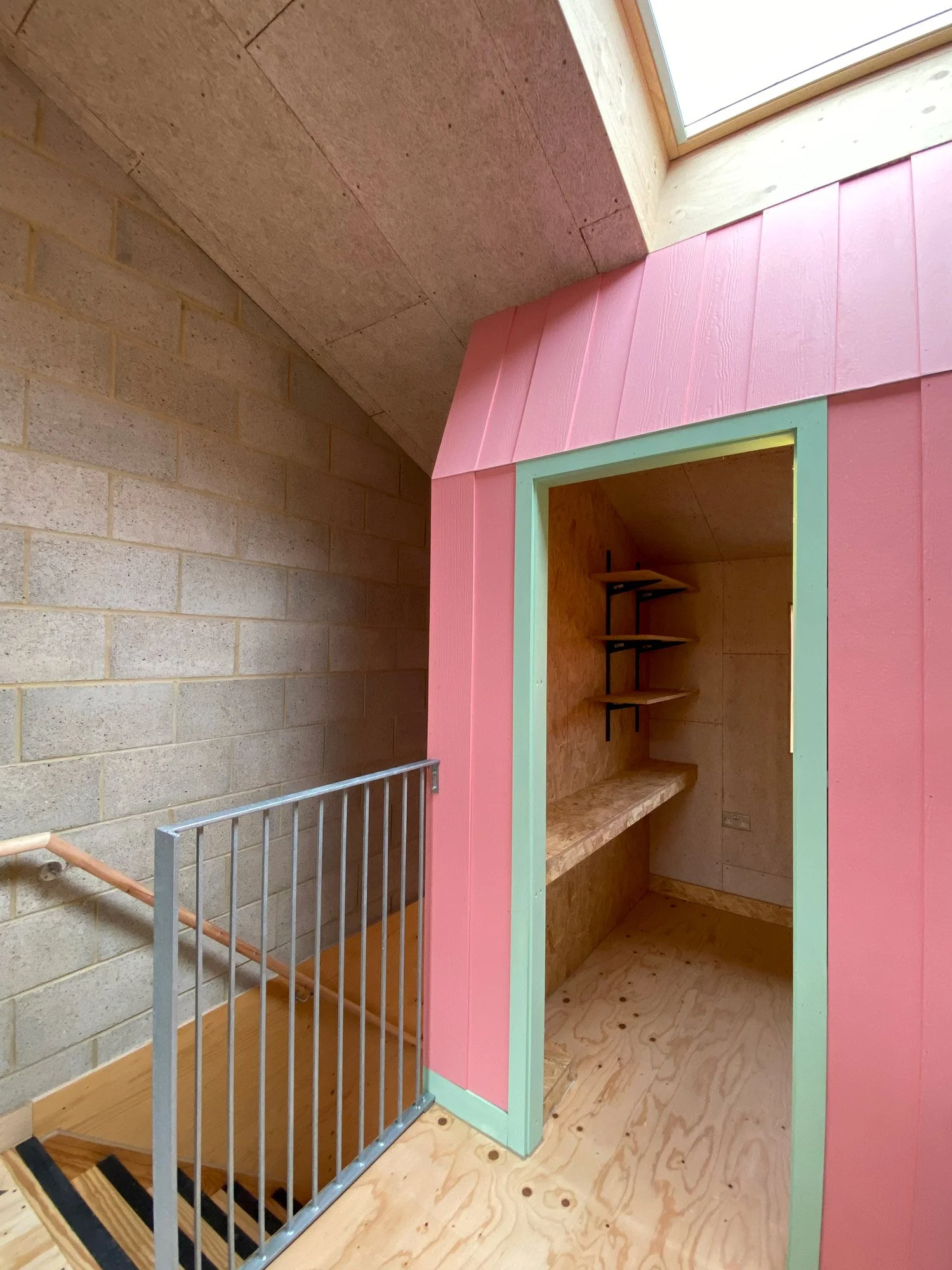 Interior of a small, colorful wooden room with pink and mint green walls, built in a corner under a skylight, with shelves and a desk inside, next to a stairway and a concrete wall.