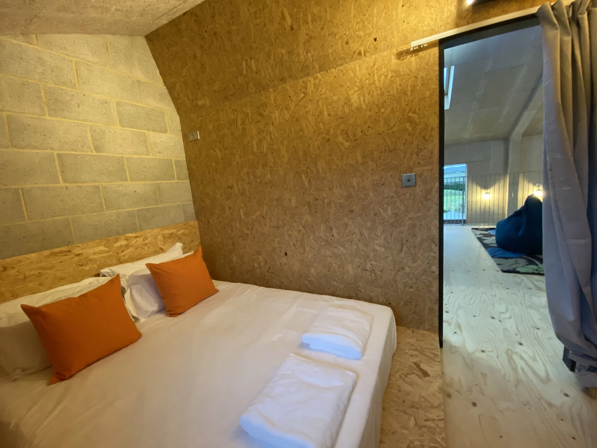 Bedroom with a bed that has white sheets and three orange pillows, wooden and concrete walls, and a doorway leading to an outdoor patio with a sliding door and railing.