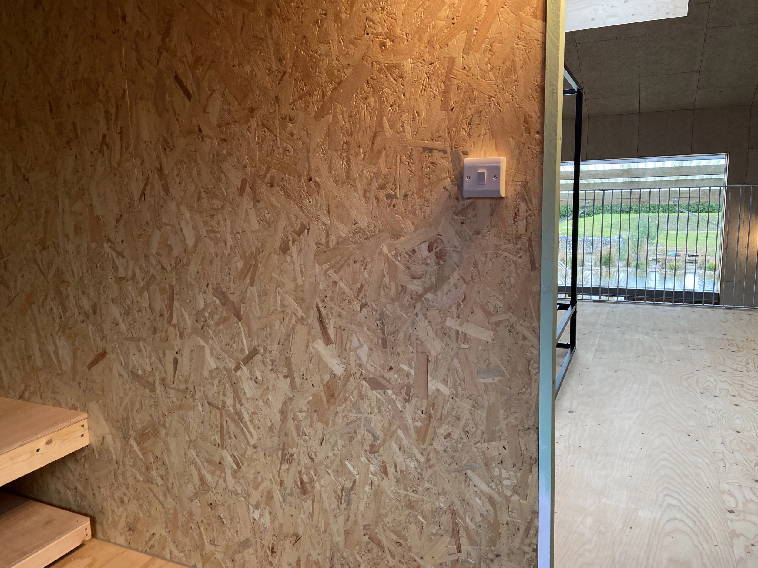 Interior view showing a partially finished wall covered in oriented strand board (OSB) with a light switch, next to a railing overlooking an outdoor landscape with grass, water, and trees.