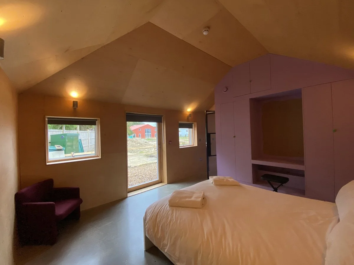 Bedroom with a bed, towels, purple built-in shelves, a window, a door, and a small purple sofa.