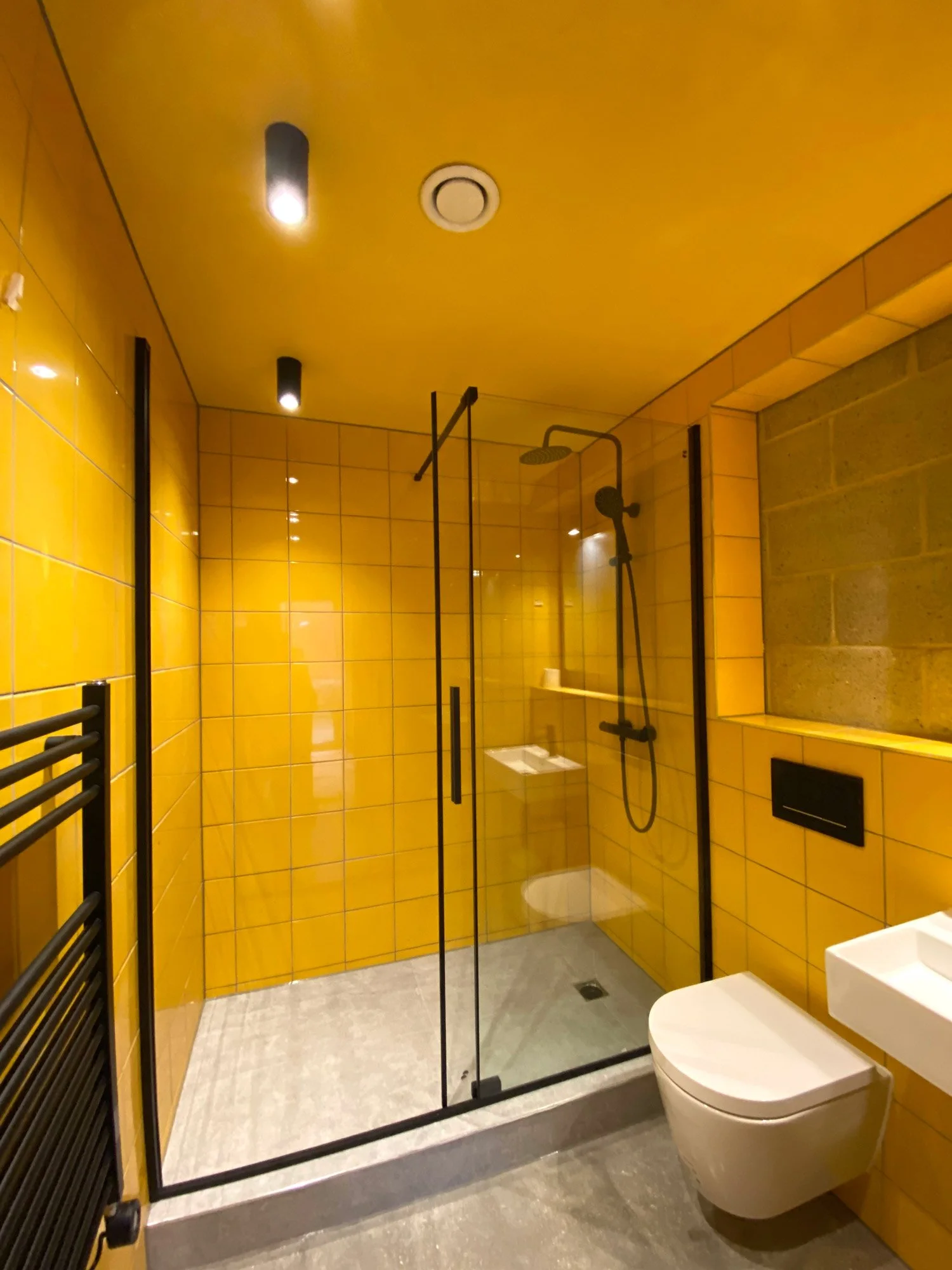 Modern bathroom with yellow tiled walls and ceiling, walk-in shower with glass door, mounted black shower head, black towel radiator, white wall-mounted toilet, small white sink, and gray tiled floor.