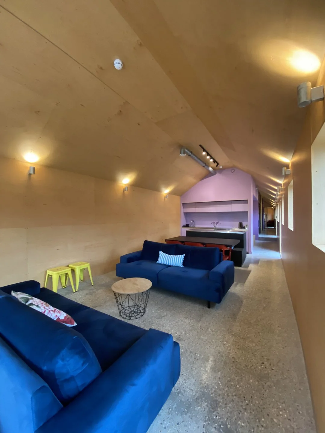 Interior of a modern living room with blue sofas, a small wooden coffee table, and colorful stools, featuring wooden panel walls and ceiling, with built-in lighting and a kitchenette area in the background.