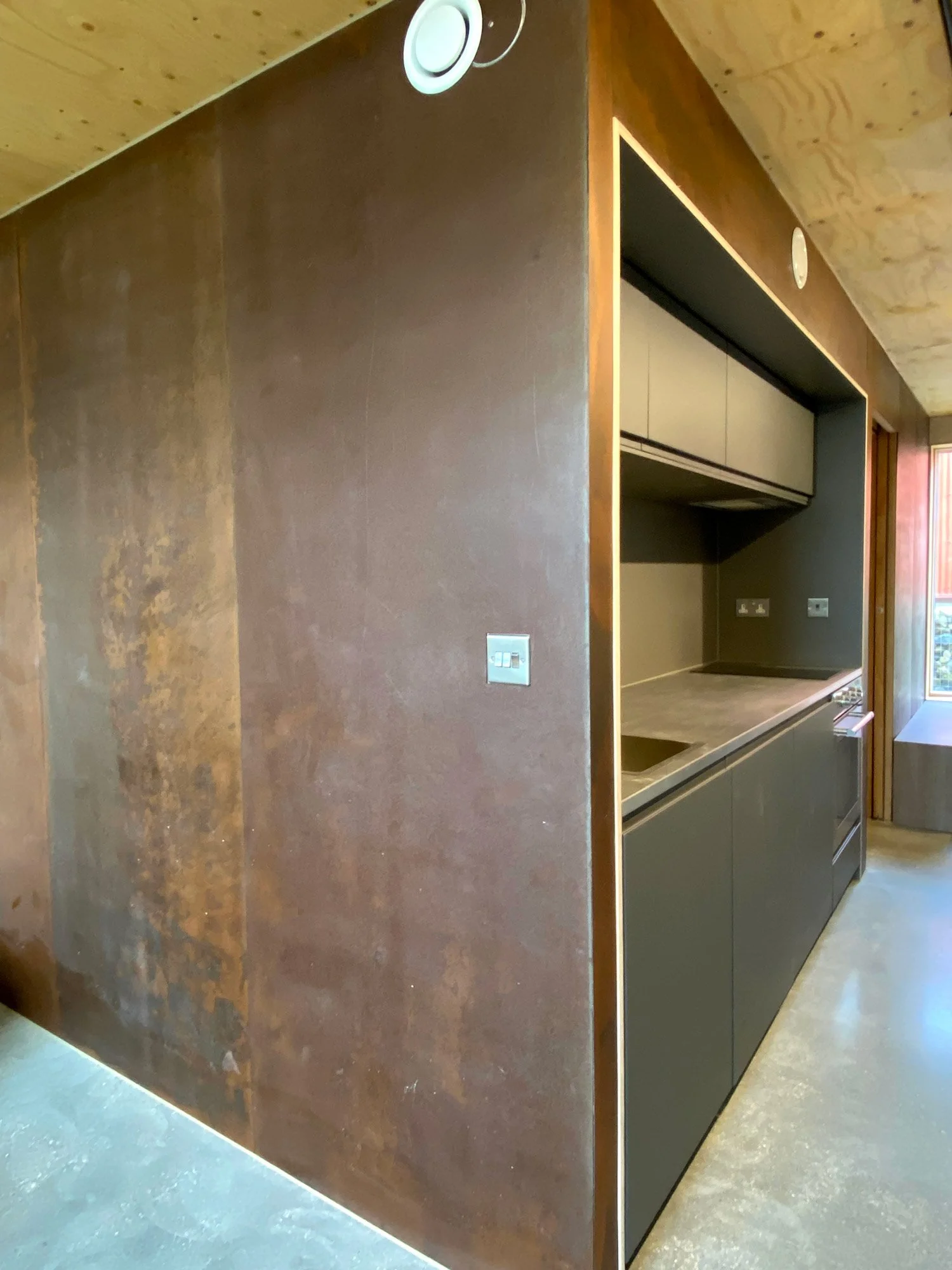 Modern kitchen with dark cabinets, a built-in oven, and a small sink, featuring a brown accent wall and a wooden ceiling.