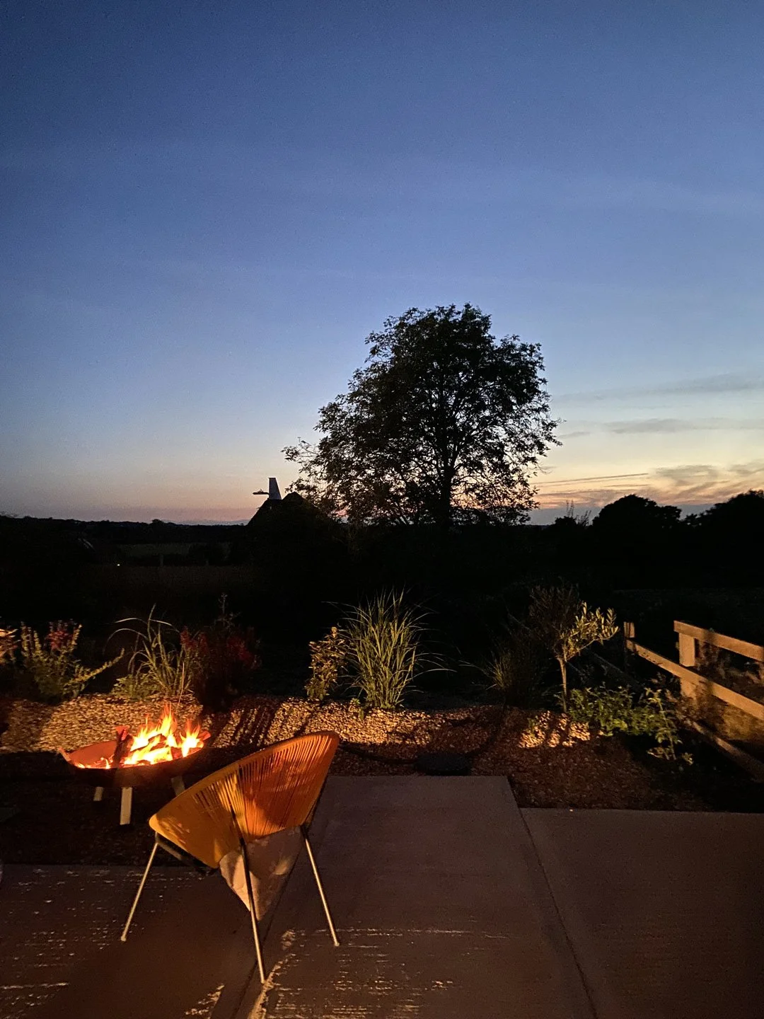 Outdoor scene at dusk with a fire pit, a modern chair, trees, and a fence against a blue and orange sky.