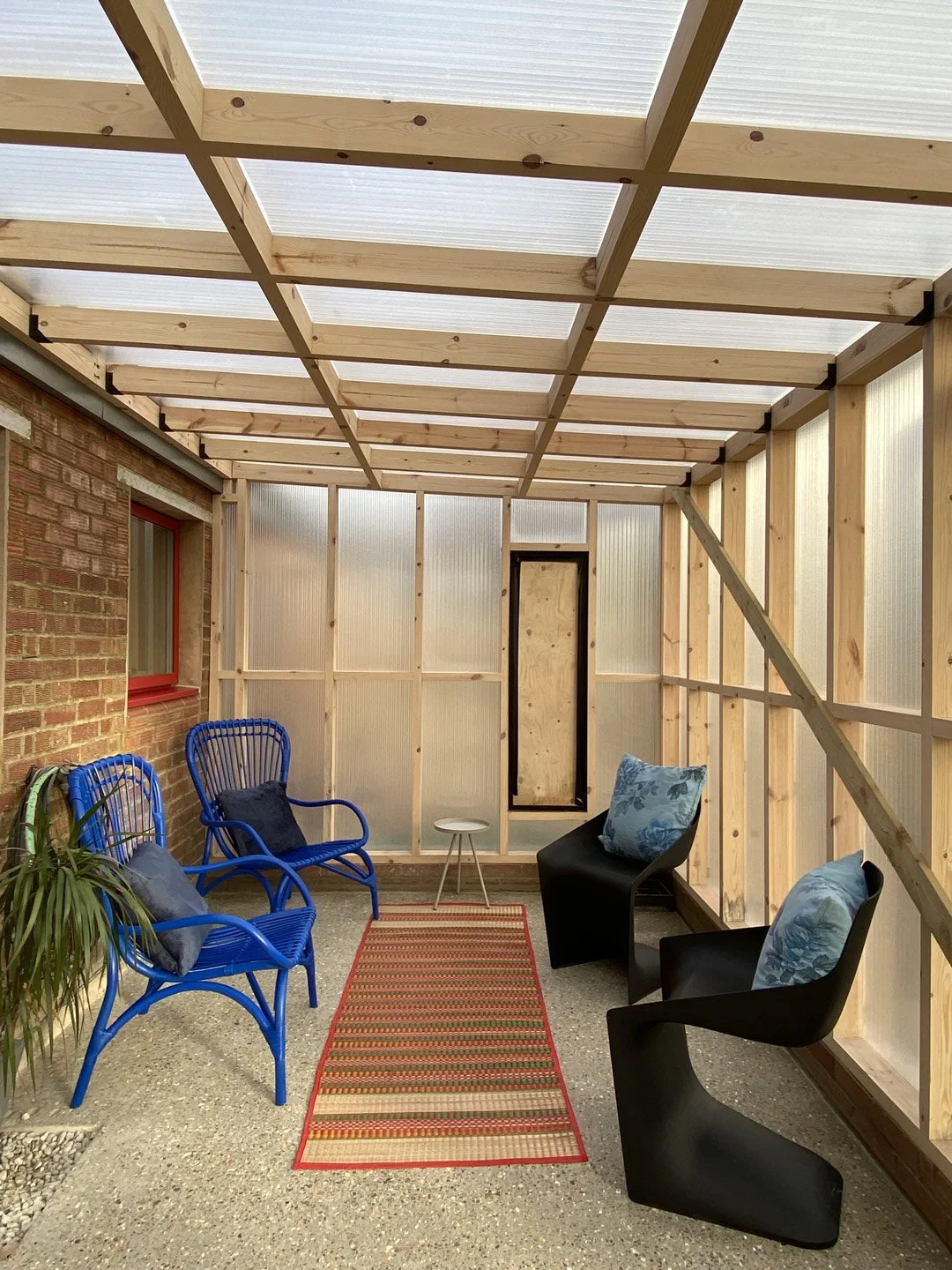 Indoor porch with unfinished wooden framing, two blue chairs with gray cushions, two black chairs with floral cushions, a small white side table, a red striped rug, a brick wall on the left, a window with red trim, and a door at the back.