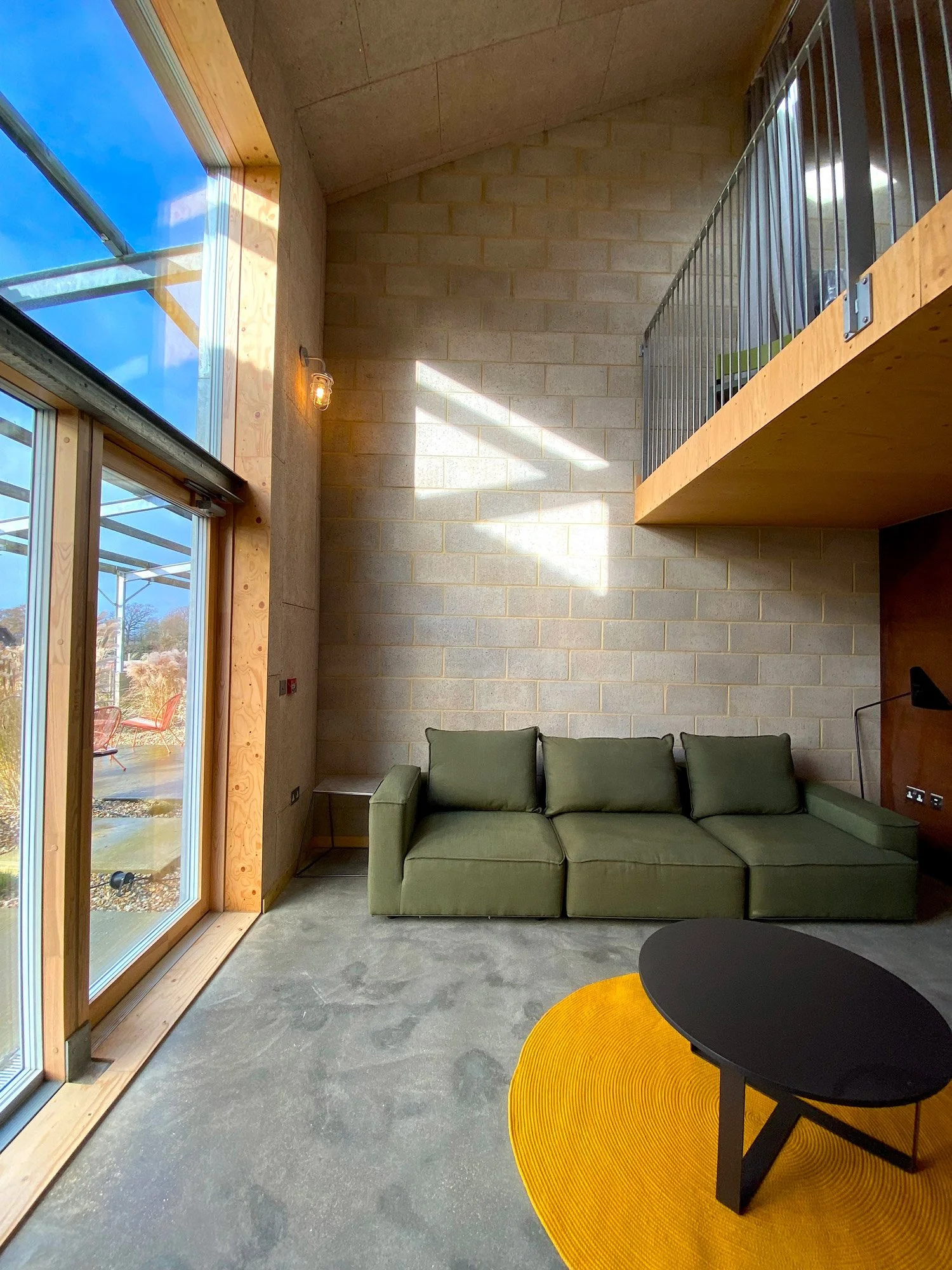 Modern living room with large windows, green sofa, black coffee table on a yellow rug, and a loft area with metal railing.
