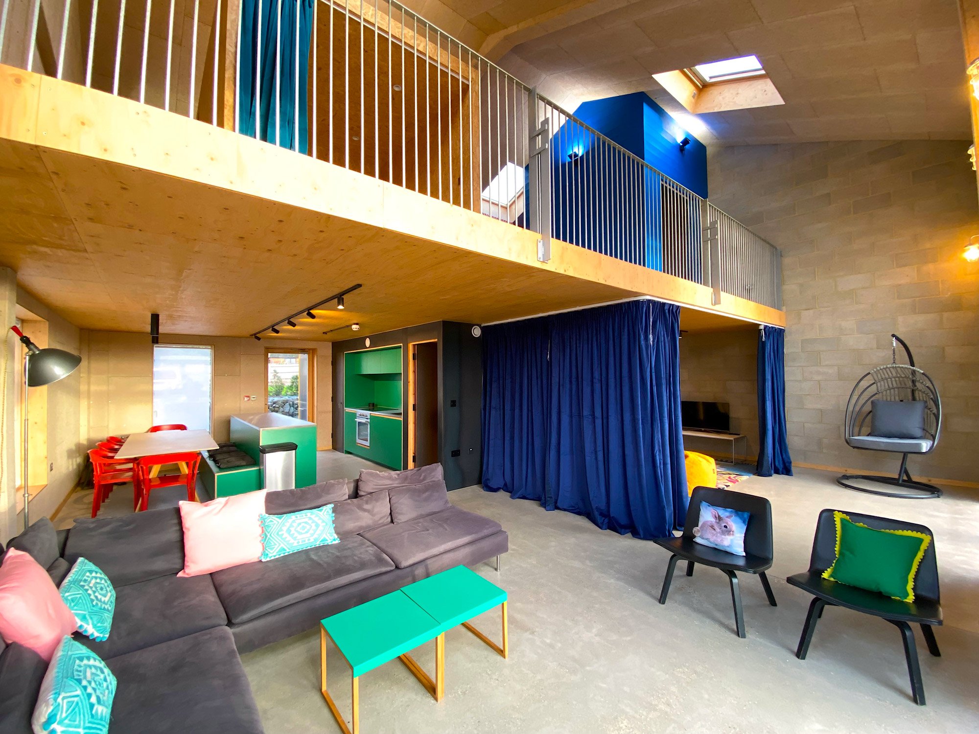 Open living room with a gray sectional sofa, colorful pillows, a teal coffee table, and seating with pillows. To the right, a hanging circular chair and a gray TV console with a TV. Behind the sofa, a blue curtain section separates the living space from a corner with a mounted TV, a yellow pouf, and toys. The upper level has a balcony with a yellow wooden railing, blue accent wall, skylights, and ceiling lights, with a blue game room and a skylight in the ceiling.