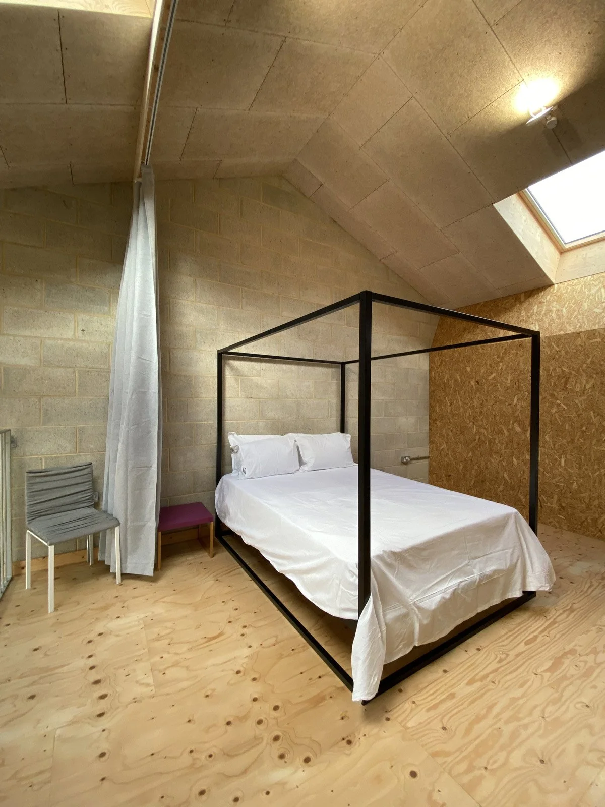 A minimalist bedroom with a black metal four-poster bed, white bedding, a small chair, a purple stool, and a window skylight in a wood-paneled ceiling.