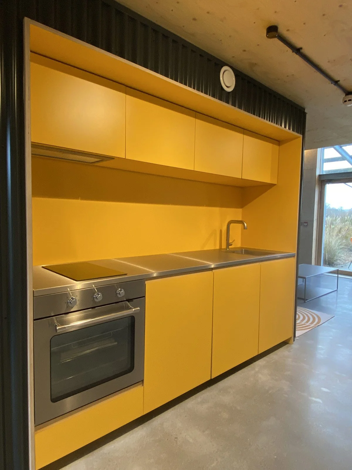 Modern kitchen with yellow cabinets, stainless steel oven, and a black faucet, situated in an industrial-style space with concrete flooring and large windows.