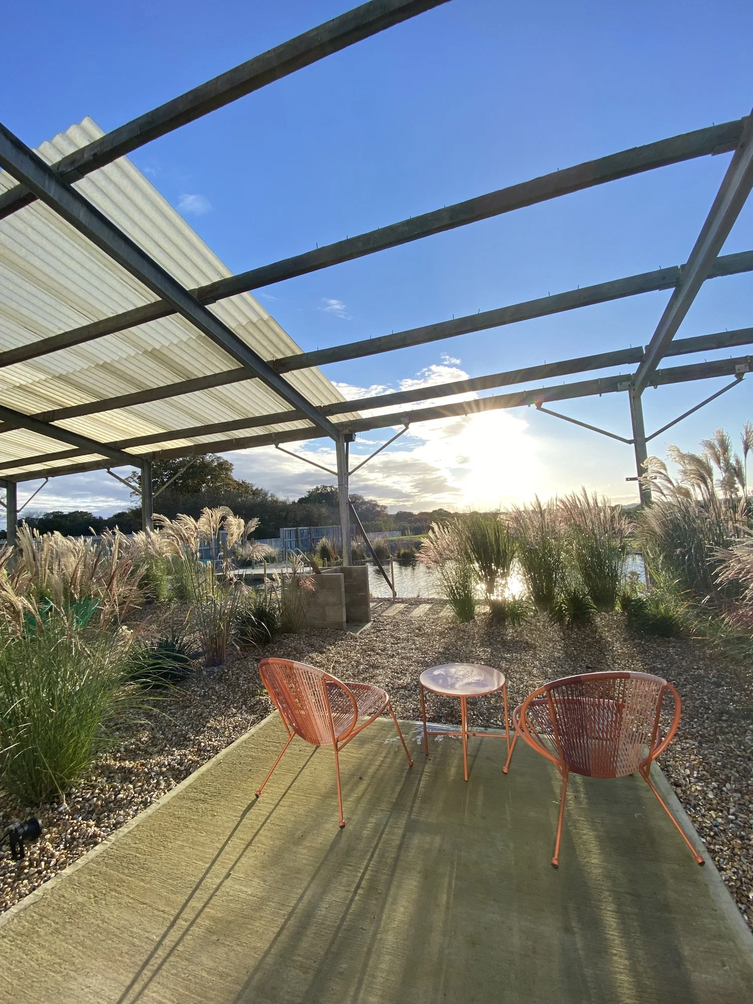 Outdoor patio with two orange chairs and a small round table on a green rug, surrounded by ornamental grasses, with a body of water and trees in the background, under a partially covered metal structure, during a sunny day.