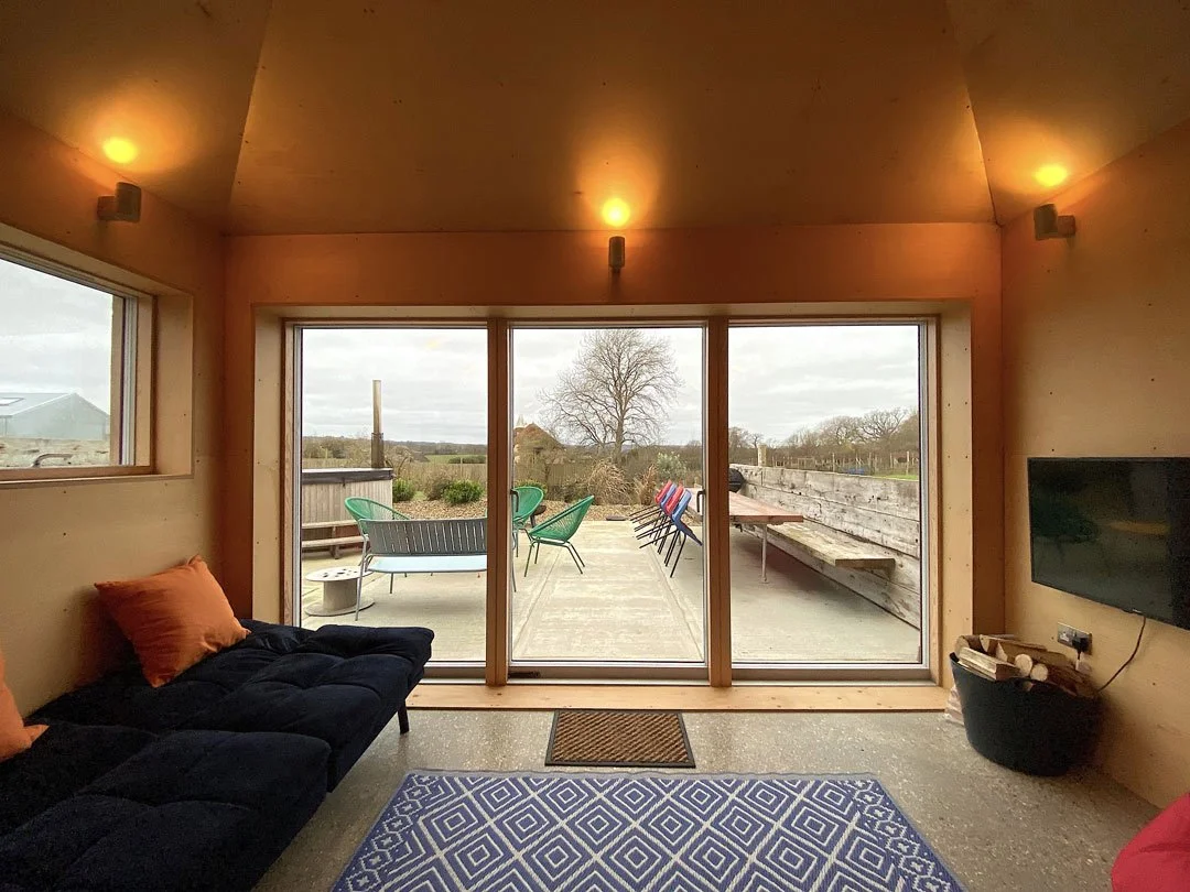 Interior of a cozy living room with a black sofa, orange pillows, a patterned blue rug, and a flat-screen TV on the wall, opening to a small outdoor patio with chairs and a view of trees and a cloudy sky.