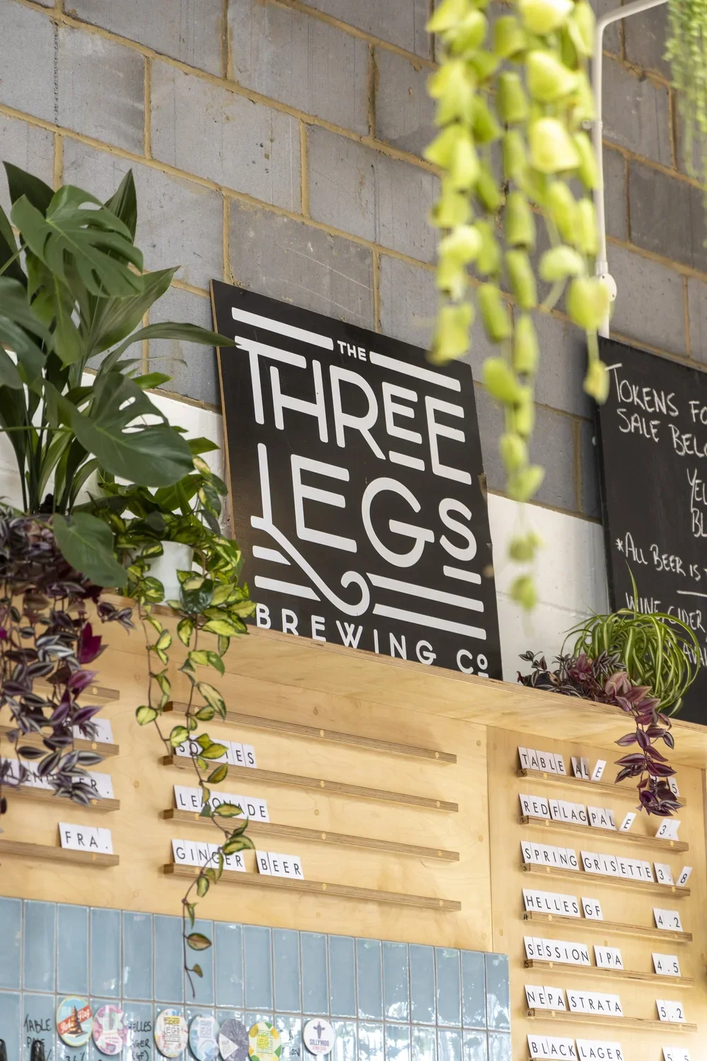 Sign for Three Legs Brewing Co. on a brick wall, surrounded by plants and chalkboards with menu items.