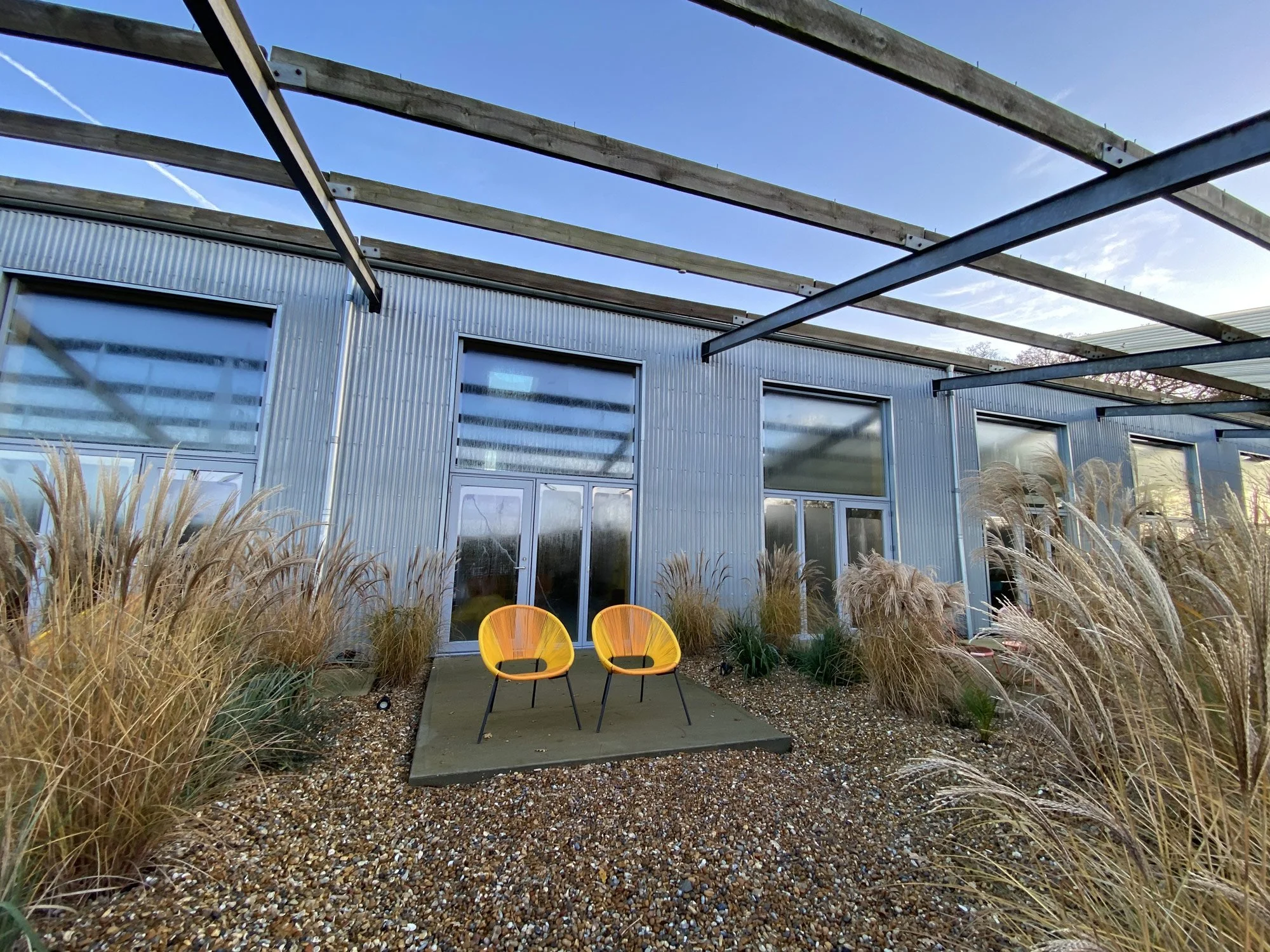 Modern building with large glass windows, yellow outdoor chairs on a small concrete patio, surrounded by ornamental grasses, under a partly cloudy sky.