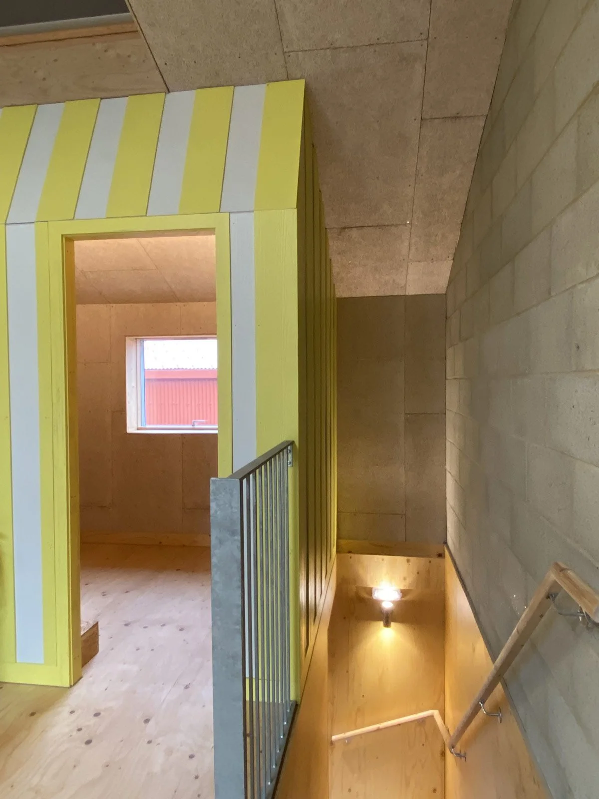 Interior of a building with a staircase, yellow and white striped small room or booth, a window, and a wooden wall.