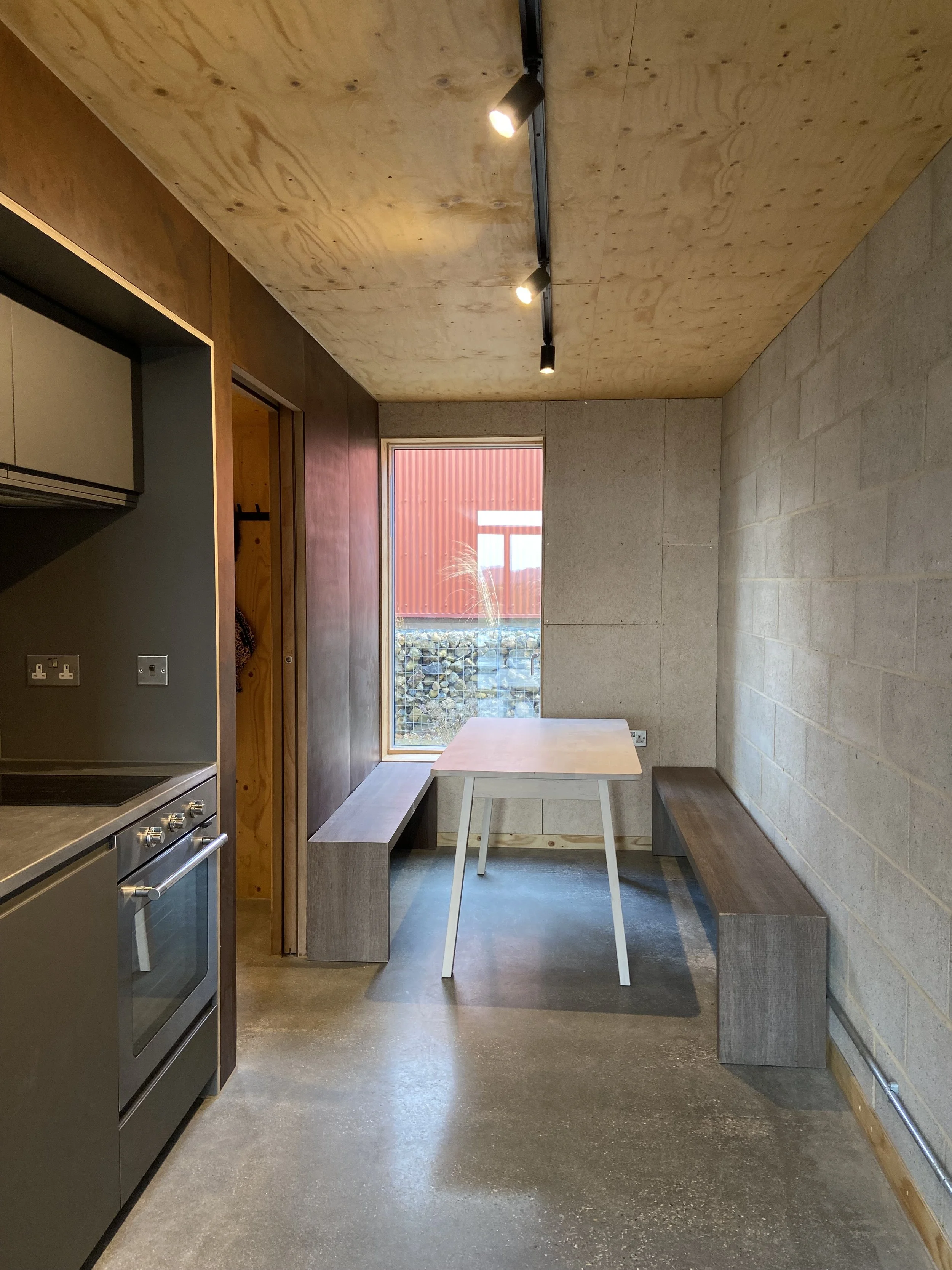 Interior of a minimalist room with a concrete floor, wooden ceiling with track lighting, a window showing a red building outside, a small white table with angled legs, a bench under the window, and a wooden wall panel.