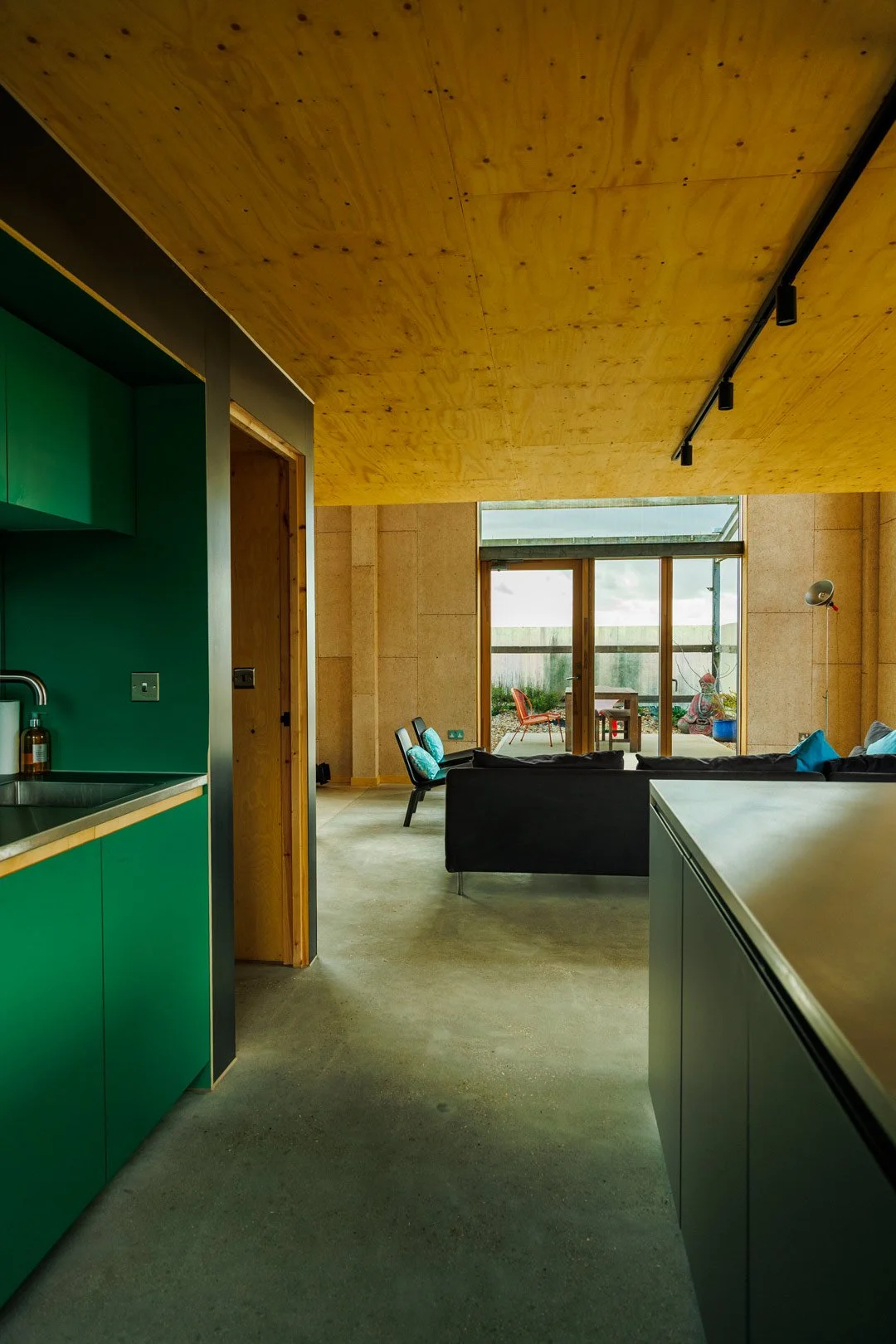 Interior view of a modern home with a kitchen on the left featuring green cabinets, and a living room with a black couch and colorful chairs, leading to a patio with outdoor furniture and garden view.