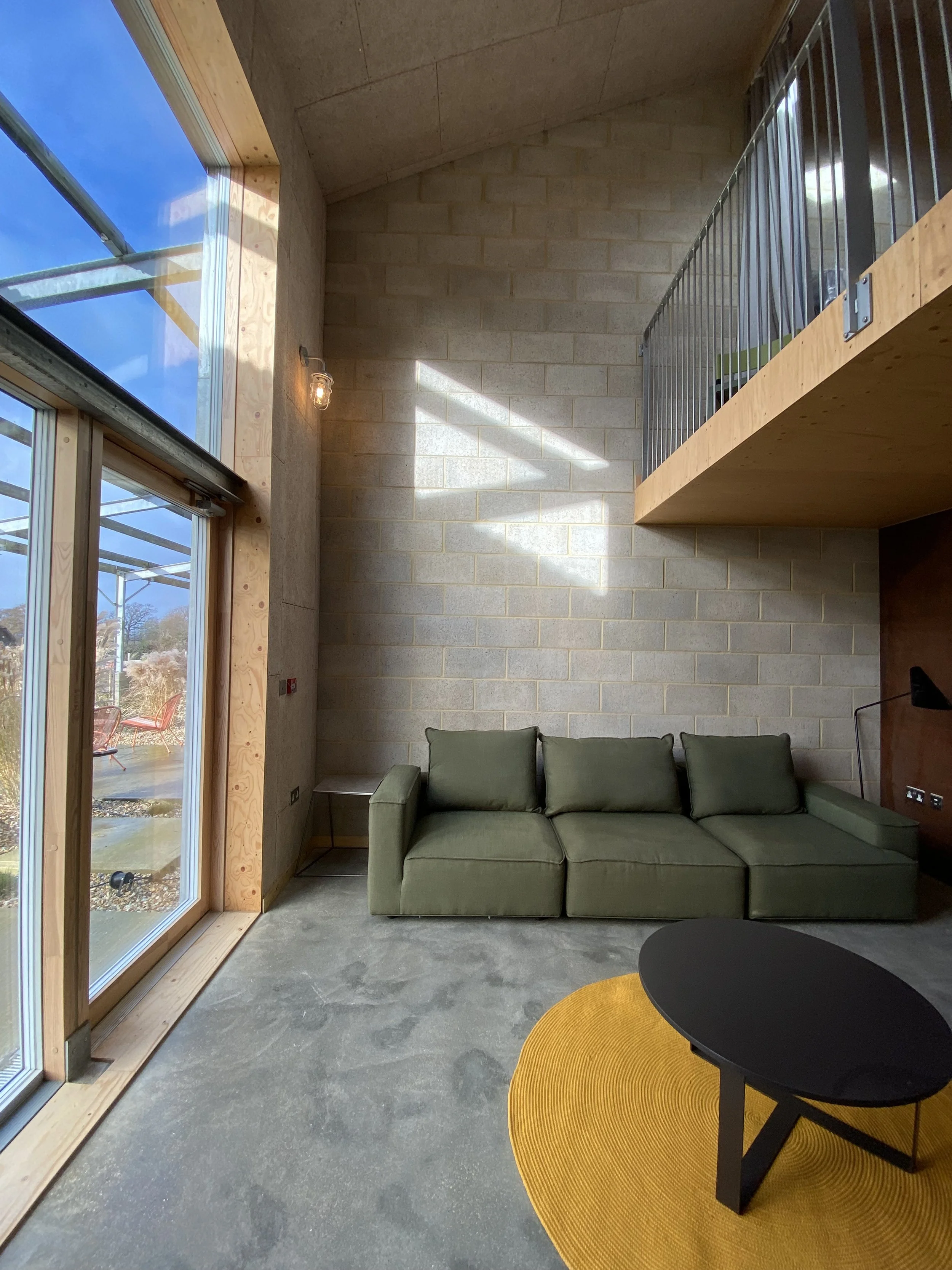 Modern living room with large floor-to-ceiling windows, a green sofa, a yellow round rug, a black coffee table, and a high ceiling with a loft and metal railing.
