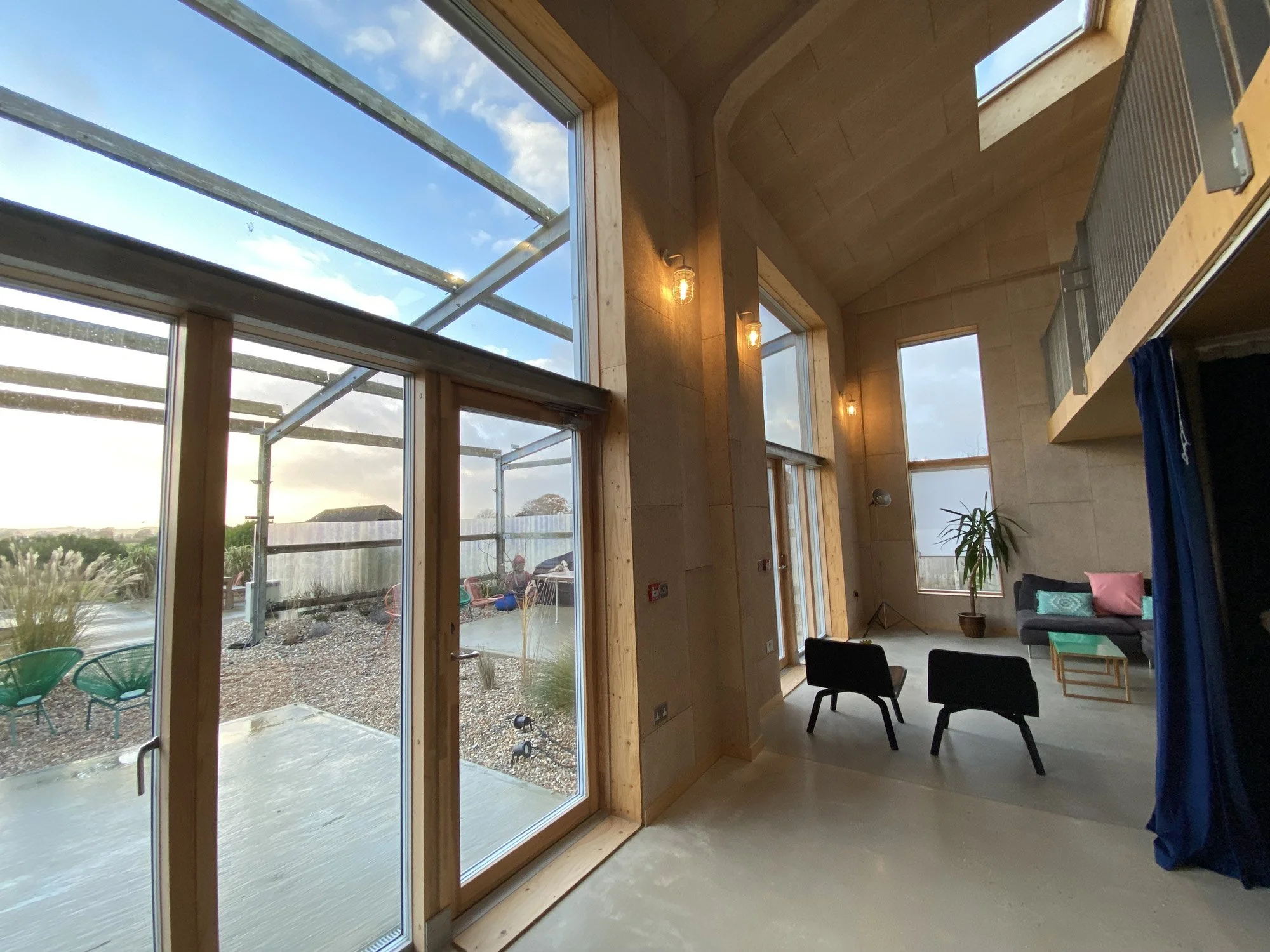 Interior view of a modern building with large windows, a seating area with black chairs and a sofa, a potted plant, and an outdoor patio visible through the windows.