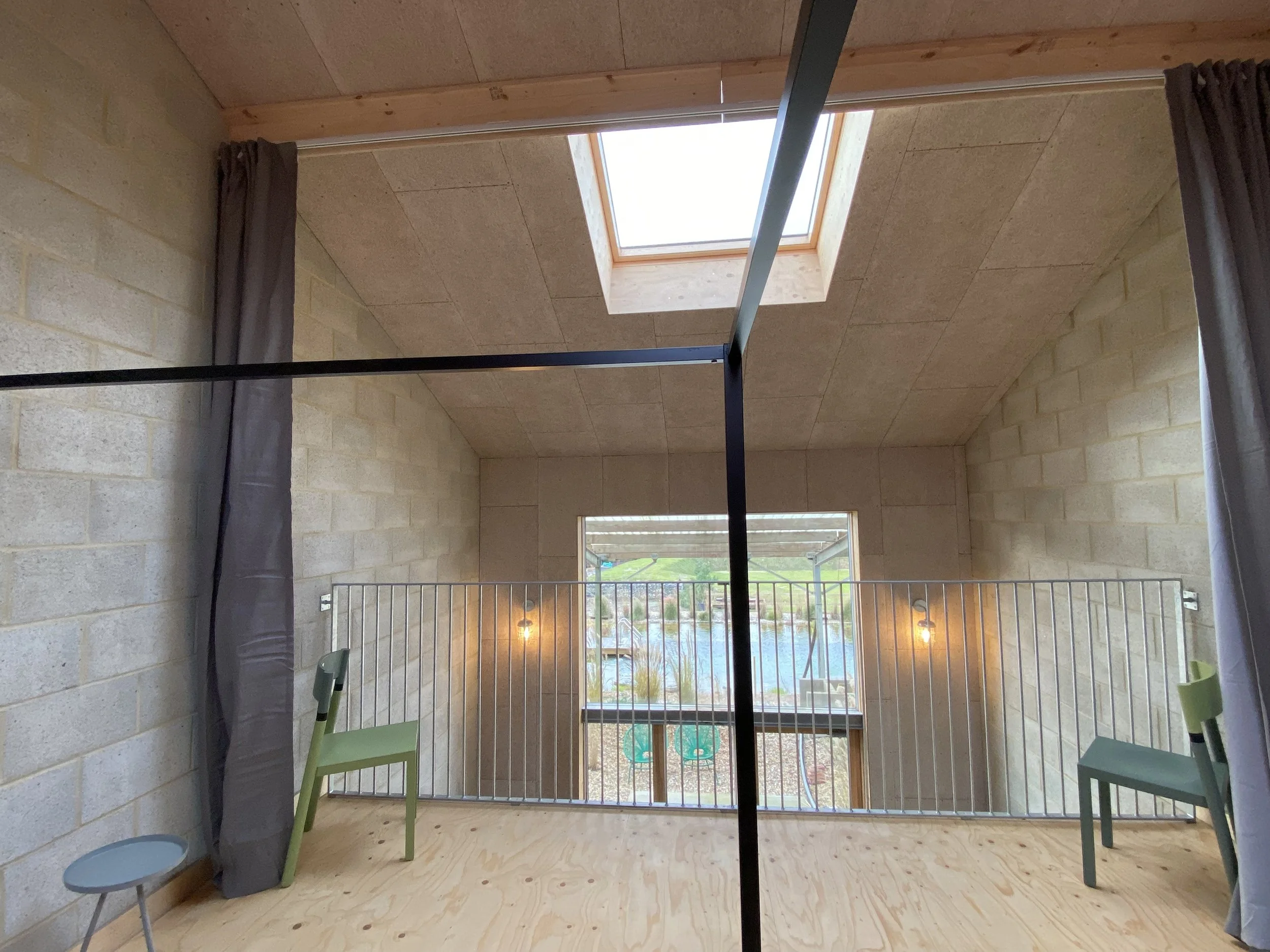 Interior of a room with a balcony view, featuring a skylight in the ceiling, two chairs, and a small table next to the window, with curtains on the sides.