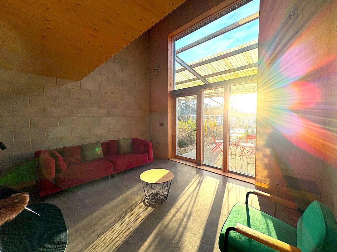 A modern living room with a red sofa, green chair, and round coffee table. Large glass sliding door leads to outdoor patio with a table and chairs. Sunlight streams in, creating a rainbow reflection on the wall.