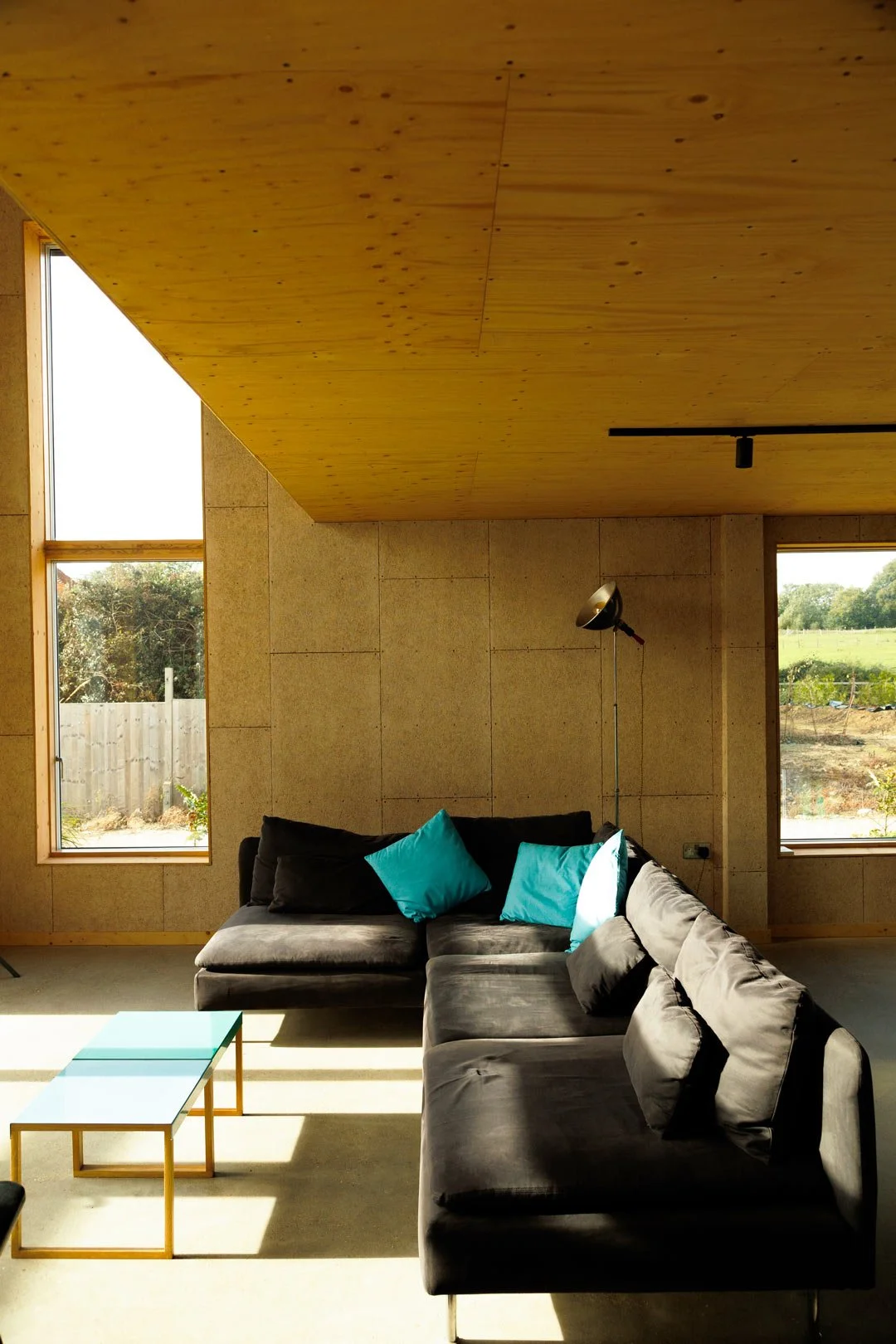 A modern living room with large windows, a black sectional sofa with teal and black pillows, a glass-top coffee table, a floor lamp, wooden ceiling and walls, and a view of a backyard with green grass and trees.