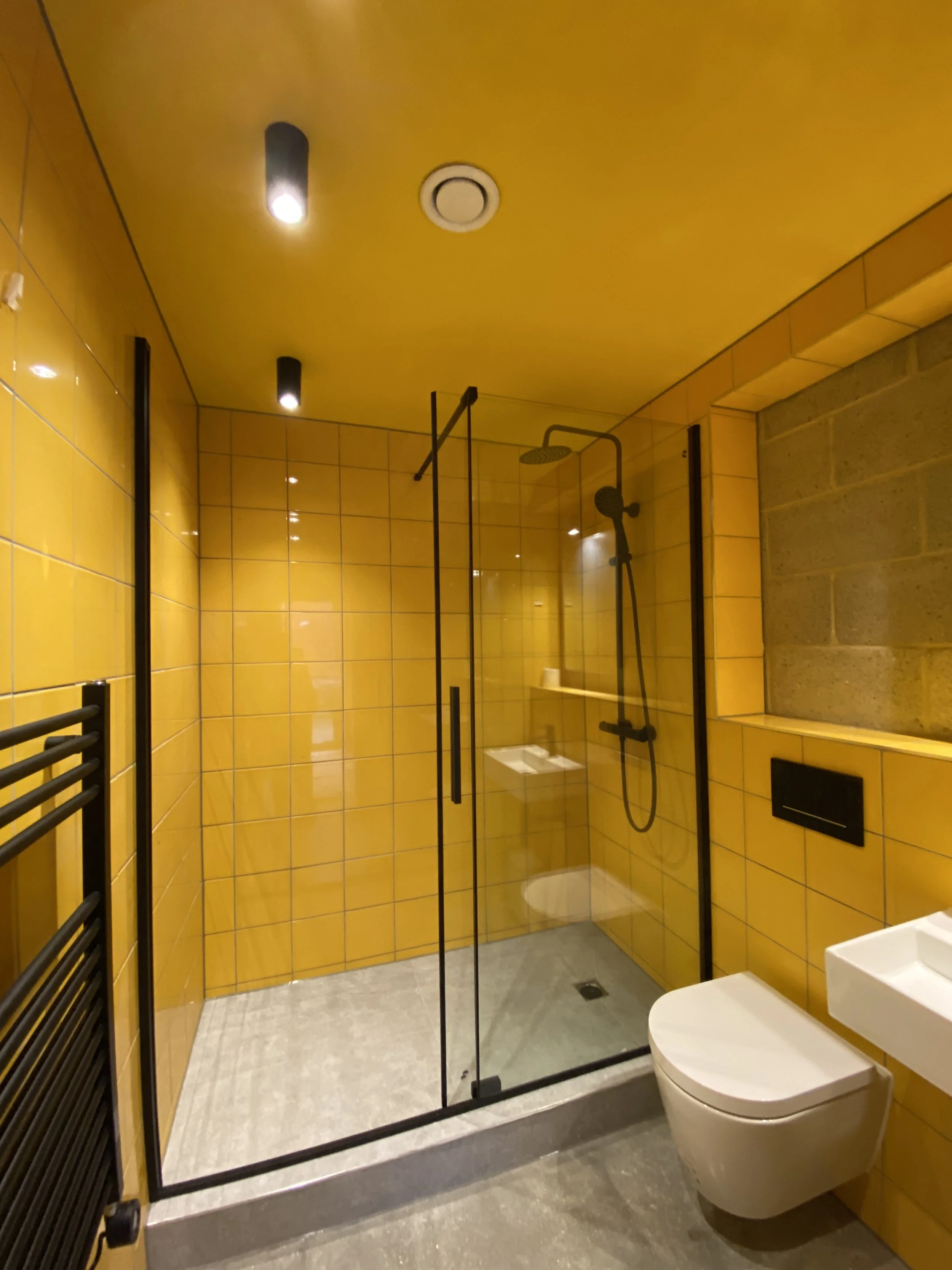 Modern bathroom with yellow tiled walls, glass shower enclosure with black fixtures, wall-mounted white toilet, and small white sink.
