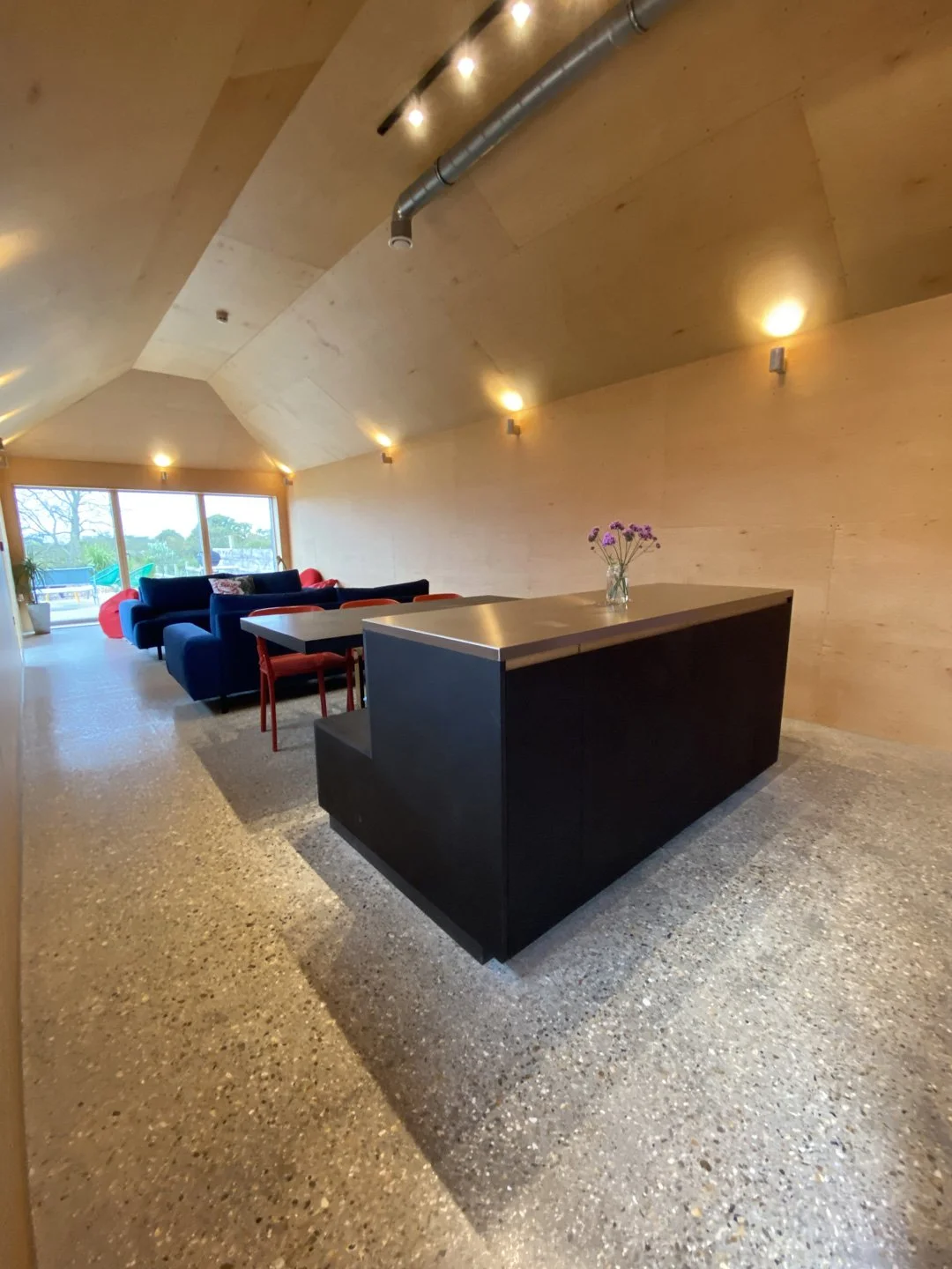 Modern living room with a black kitchen island, a vase of purple flowers, a row of black and red sofas, and large windows overlooking a backyard.