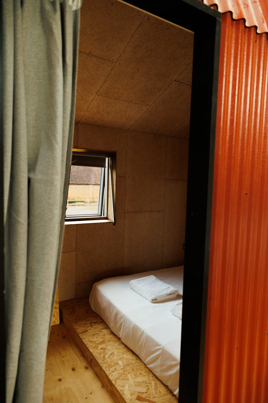 Small bedroom with wooden wall panels, a rectangular window, a bed with folded towels, and a curtain partially pulled aside.