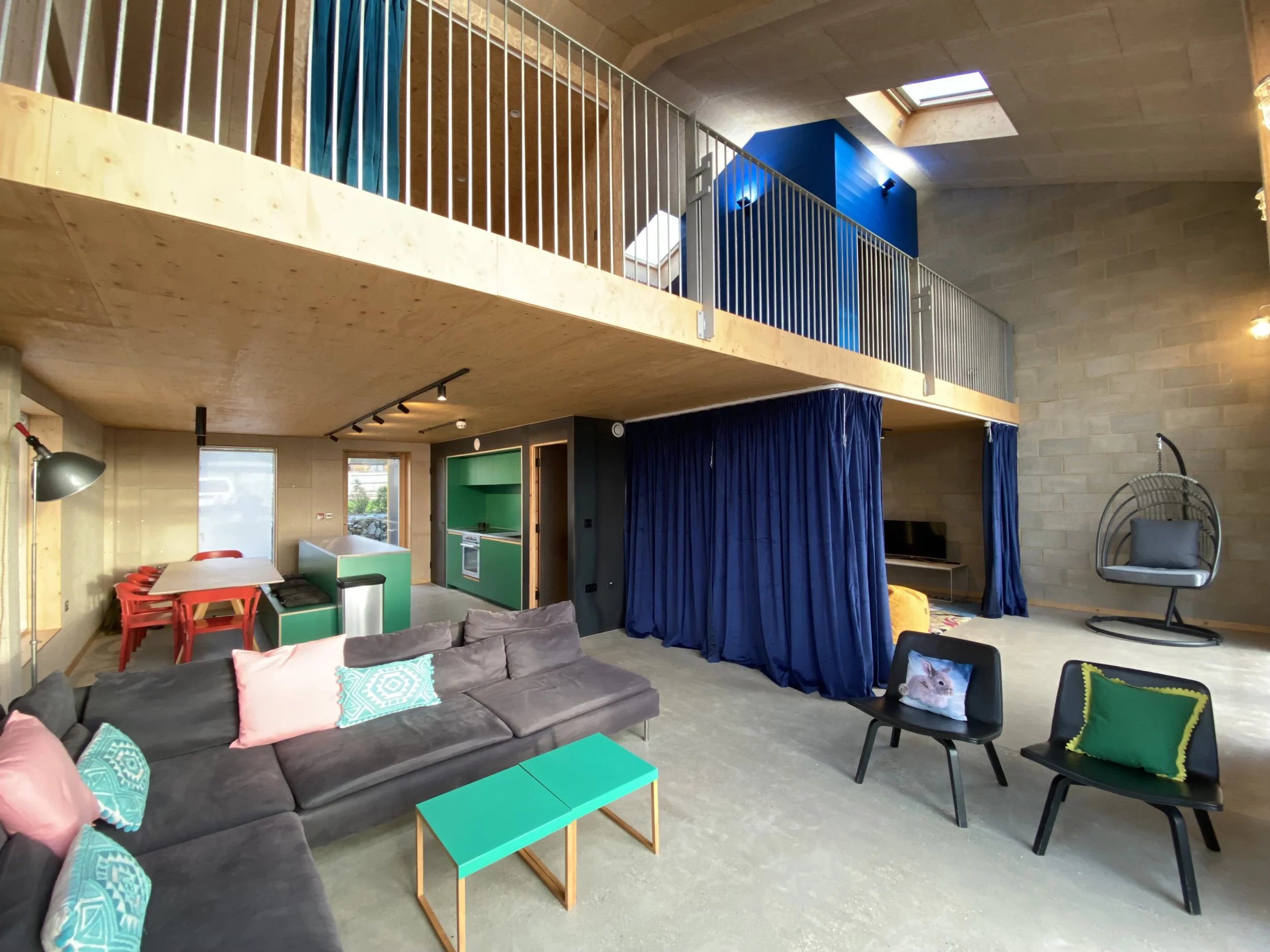 Open-concept living space with a grey sofa and turquoise pillows, a turquoise coffee table, and black chairs with colorful pillows. The room has a kitchen area with a green and wood kitchen set, a dining table with red chairs, and a built-in blue cur