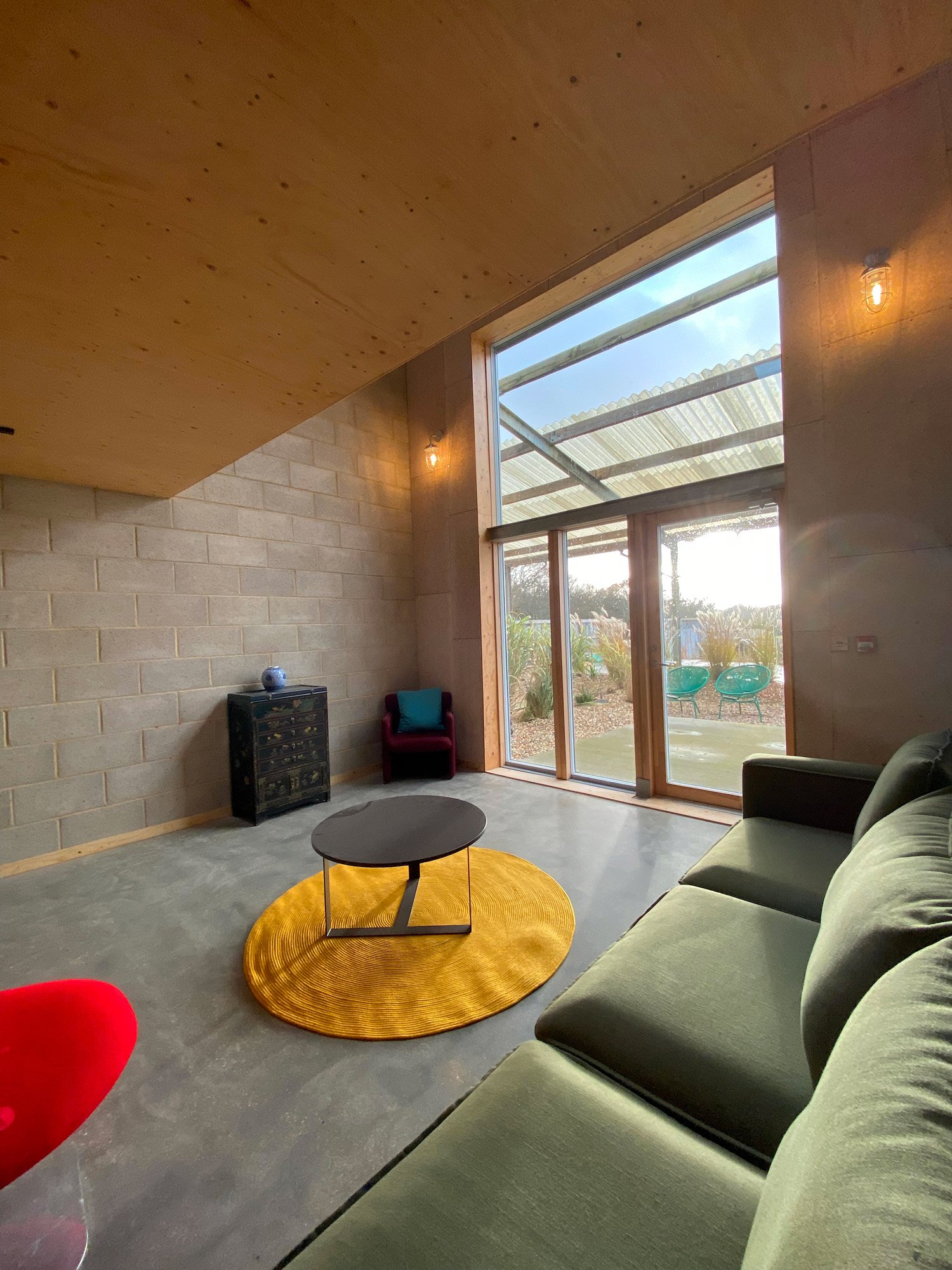 Living room with high ceiling, large window, green sofa, small round table on a yellow rug, and outdoor plants visible through window.