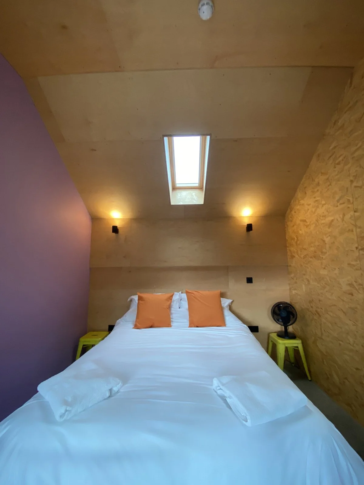 A bedroom with a bed, two orange pillows, and yellow side tables. There is a skylight in the wooden ceiling, and a small fan on a yellow stool next to one of the side tables.