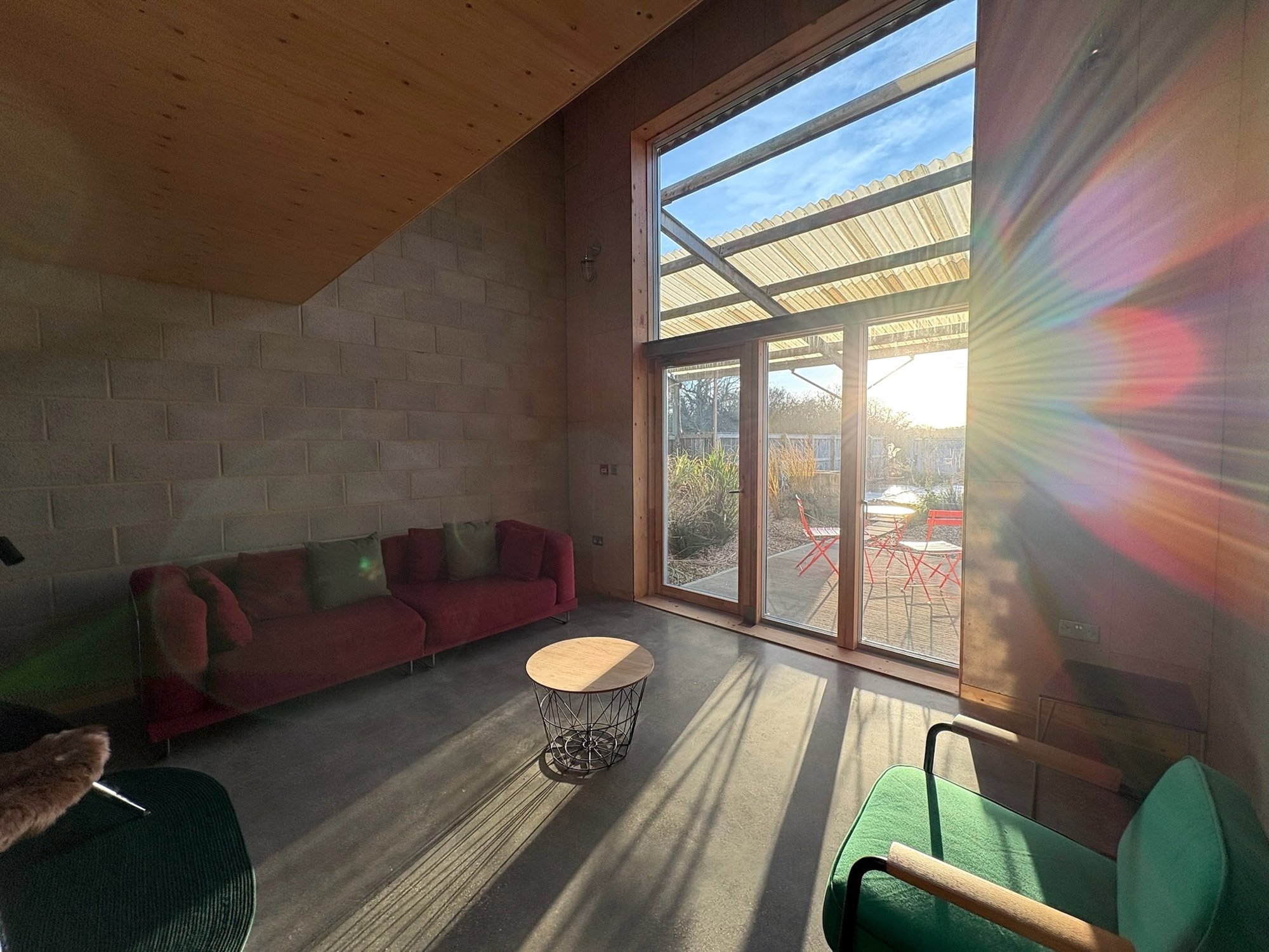 Sunlight streaming into a living room with a red sofa, green pillows, a round coffee table, a green armchair, and a large window revealing an outdoor patio with red chairs and a table. Sun rays create a glare and rainbow effect.