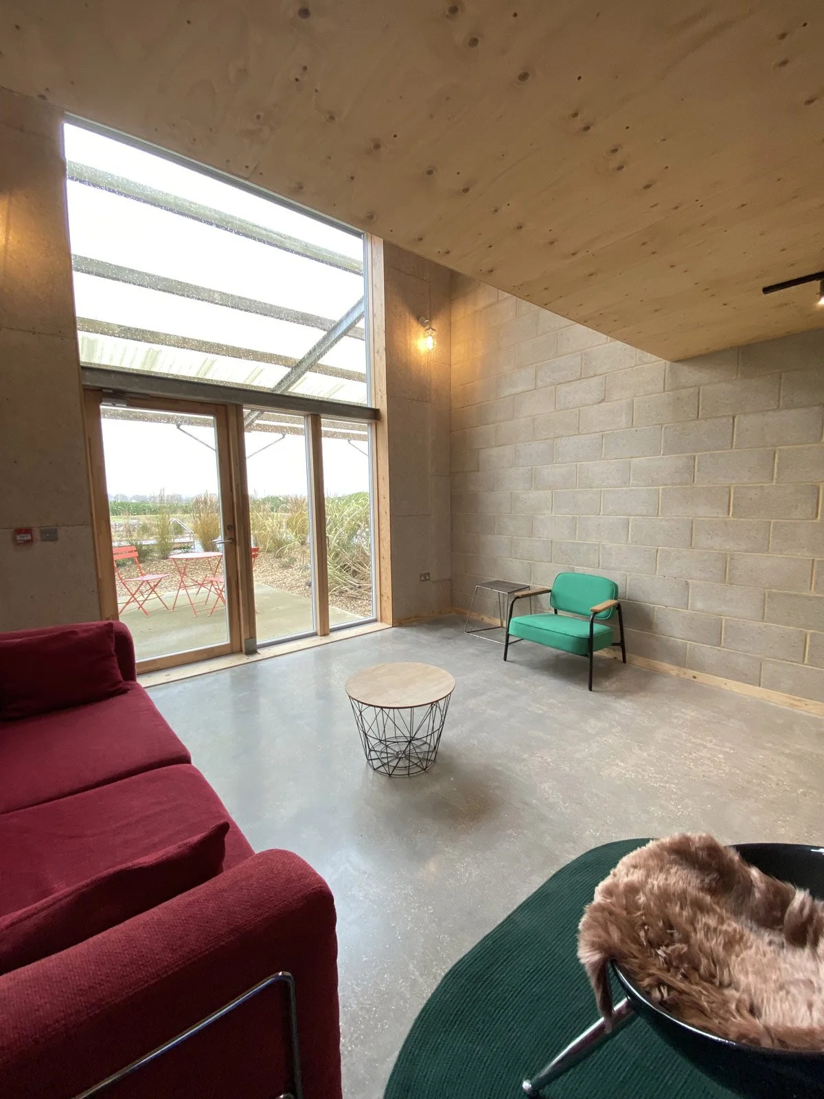 Interior of a minimalist living room with red and green sofas, a round coffee table, and large glass doors leading outside to a patio with outdoor furniture and plants.