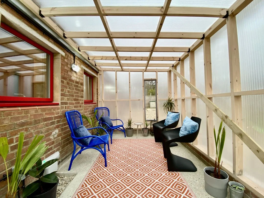 A partially enclosed sunroom with unfinished wooden framing, a brick wall with red window frames on the left, blue and black chairs with cushions, potted plants, a patterned rug, and a transparent roof.