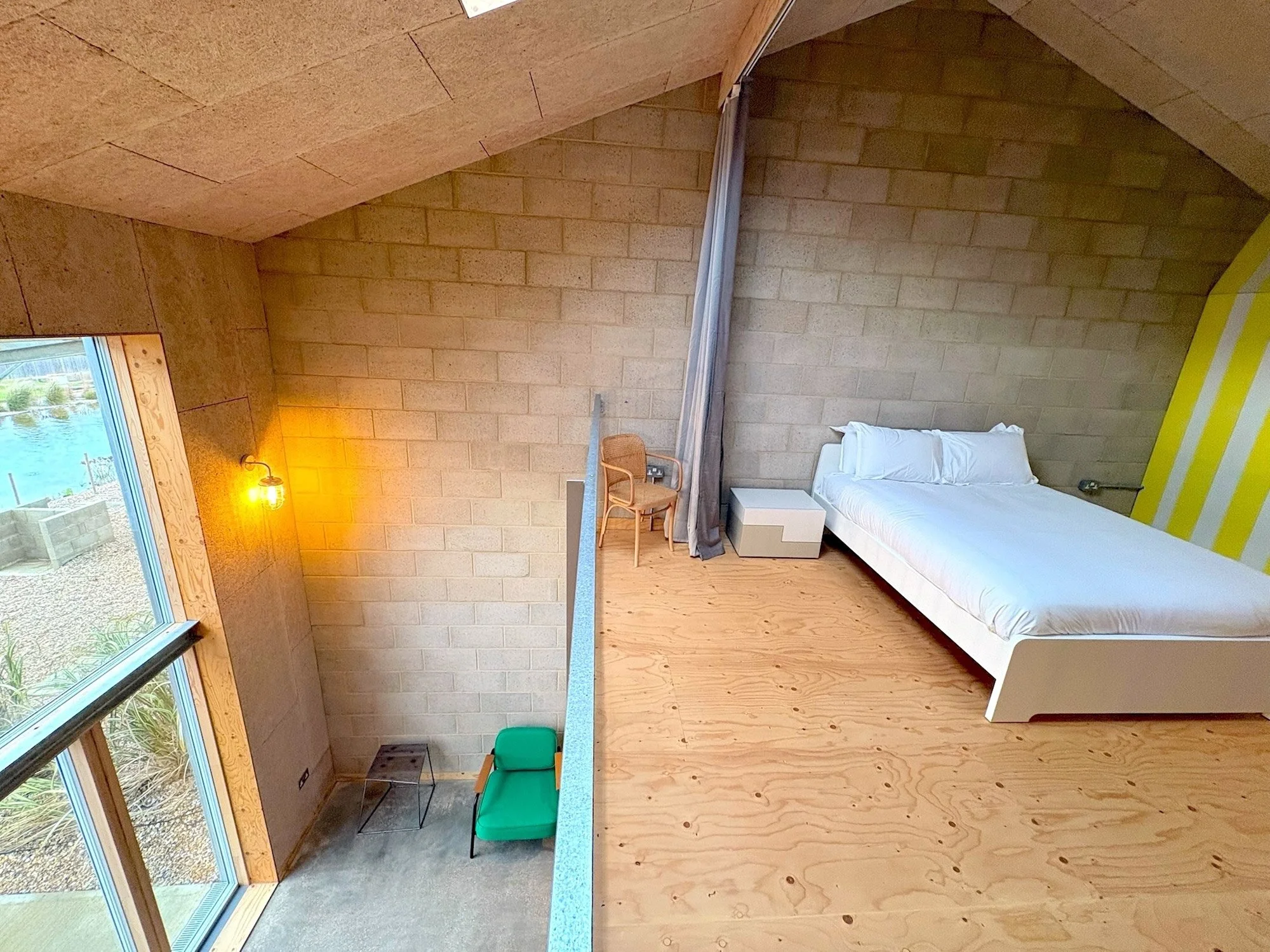 A modern bedroom with a white bed, small nightstand, wicker chair, gray curtain, wooden floor, brick wall, large window, and yellow striped wall panel.
