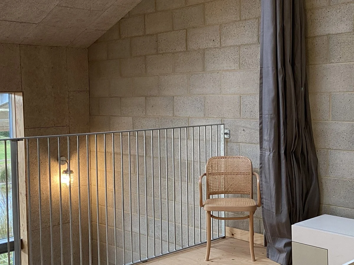 Interior of a room with beige brick walls, a metal railing, a beige woven chair, a dark curtain, and a white drawer, with a window showing outdoor scenery.