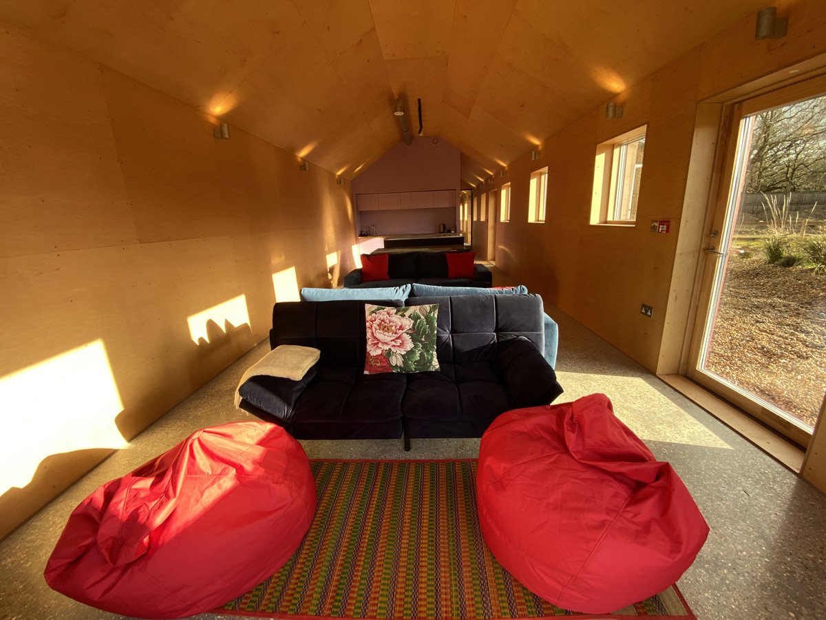 Interior of a cozy wooden room with a row of windows, showing a view of a garden outside. The room has a black couch with floral and solid cushions, two red bean bag chairs, a colorful rug, and a kitchenette in the background.
